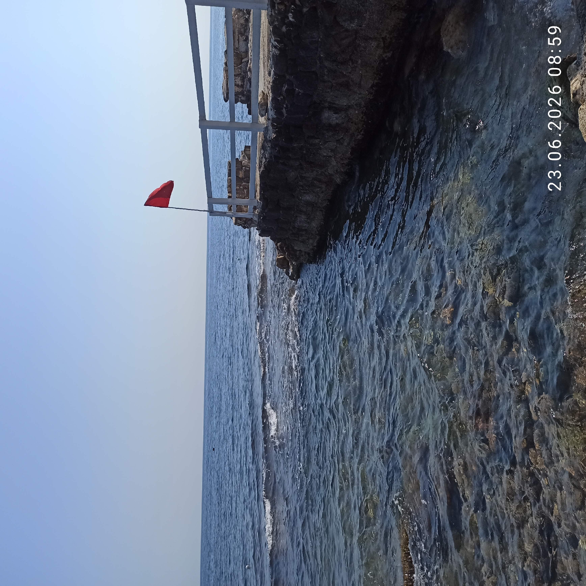 The image captures a serene coastal scene with the clear blue sea meeting a rocky shoreline. A white railing runs along the top of the rocky edge, suggesting a walkway or viewing platform. A bright red flag is prominently displayed, attached to a pole extending from the railing, likely indicating a warning or advisory for swimmers. The water is calm, with gentle waves lapping against the shore. Sunlight reflects off the water's surface, hinting at a clear day. The timestamp in the upper right corner indicates the image was taken on June 23, 2026, at 8:59 AM, suggesting it's early morning. The presence of the flag and the calm sea points towards a potentially safe but possibly regulated area for water activities. The rocky terrain and the railing suggest a developed coastal area, possibly a public beach or a scenic overlook.