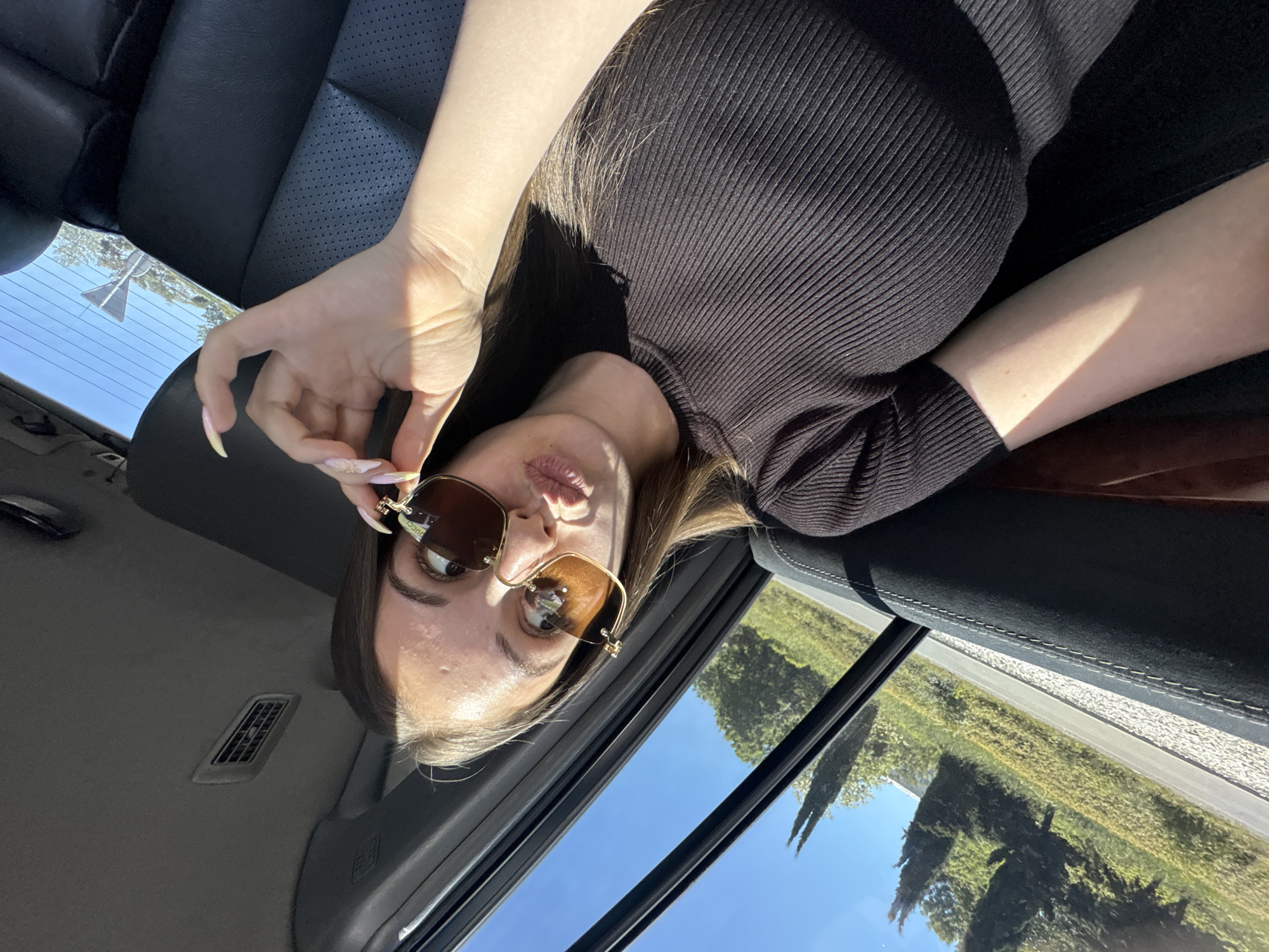 A selfie-style, canted-angle photograph taken inside a car. A woman with fair skin, brown hair, and Gucci sunglasses is the primary subject. She is wearing a ribbed, dark-colored shirt. Part of the car's interior is visible, including the leather seats and door panel. Outside the window, trees and a blue sky are visible. No indication of Burgas, Bulgaria.