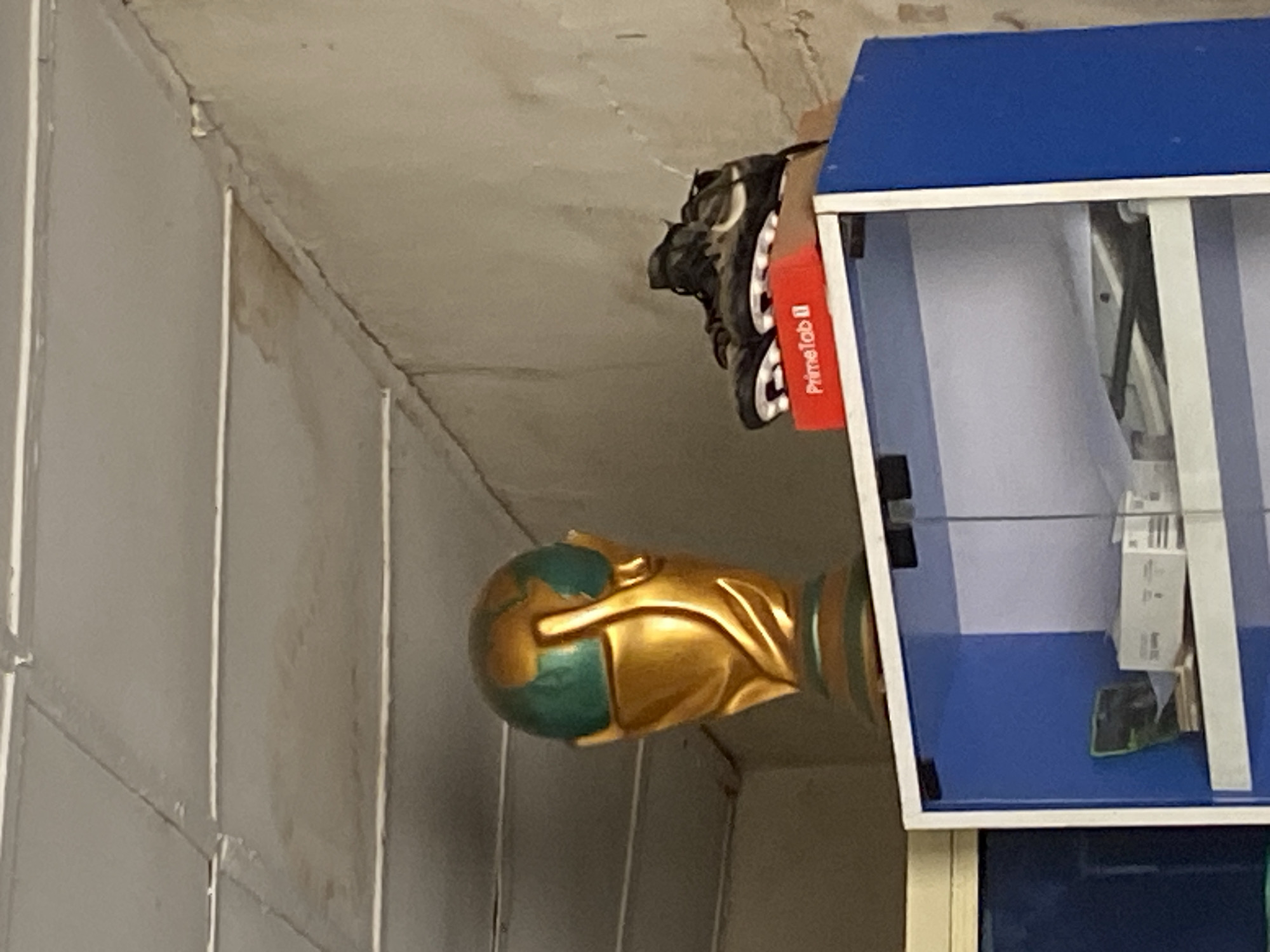 The image depicts a trophy resembling the FIFA World Cup, a pair of soccer cleats placed on a red box, and shelves containing various items. The trophy is gold and green, with the globe prominently displayed. The cleats are dark in color with white accents and cleats on the sole. The red box is labeled "PrimeTab II". The shelves are part of a blue and white cabinet. On the shelves, there are white boxes and what appears to be a green object. The scene appears to be indoors, possibly in a room in Bukuru, Nigeria, given the context. The ceiling is paneled and shows some staining. The walls are rough and textured. The lighting suggests an indoor environment. There are no people visible in the image.