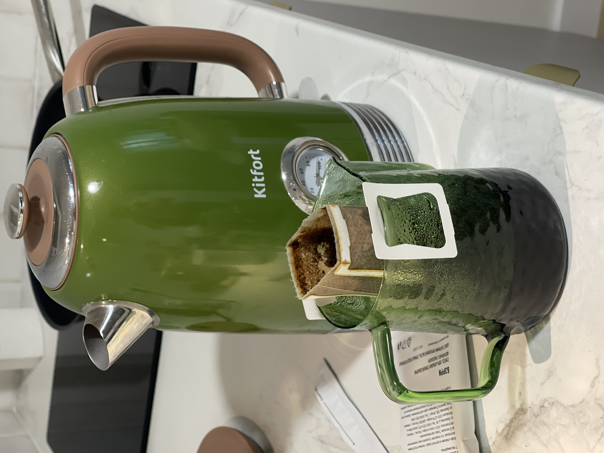 A green Kitfort electric kettle with a brown handle stands on a white countertop next to a green glass mug with a coffee bag brewing inside. The mug has condensation on the exterior. The countertop is a light marble pattern. A packet of "BARRY" brand coffee, with product information in Russian, sits beneath the mug. The OCR text on the coffee packet includes the phrases "наслаждайтесь", "изготовитель", and "Баранова", a street name in Izhevsk, Russia.
