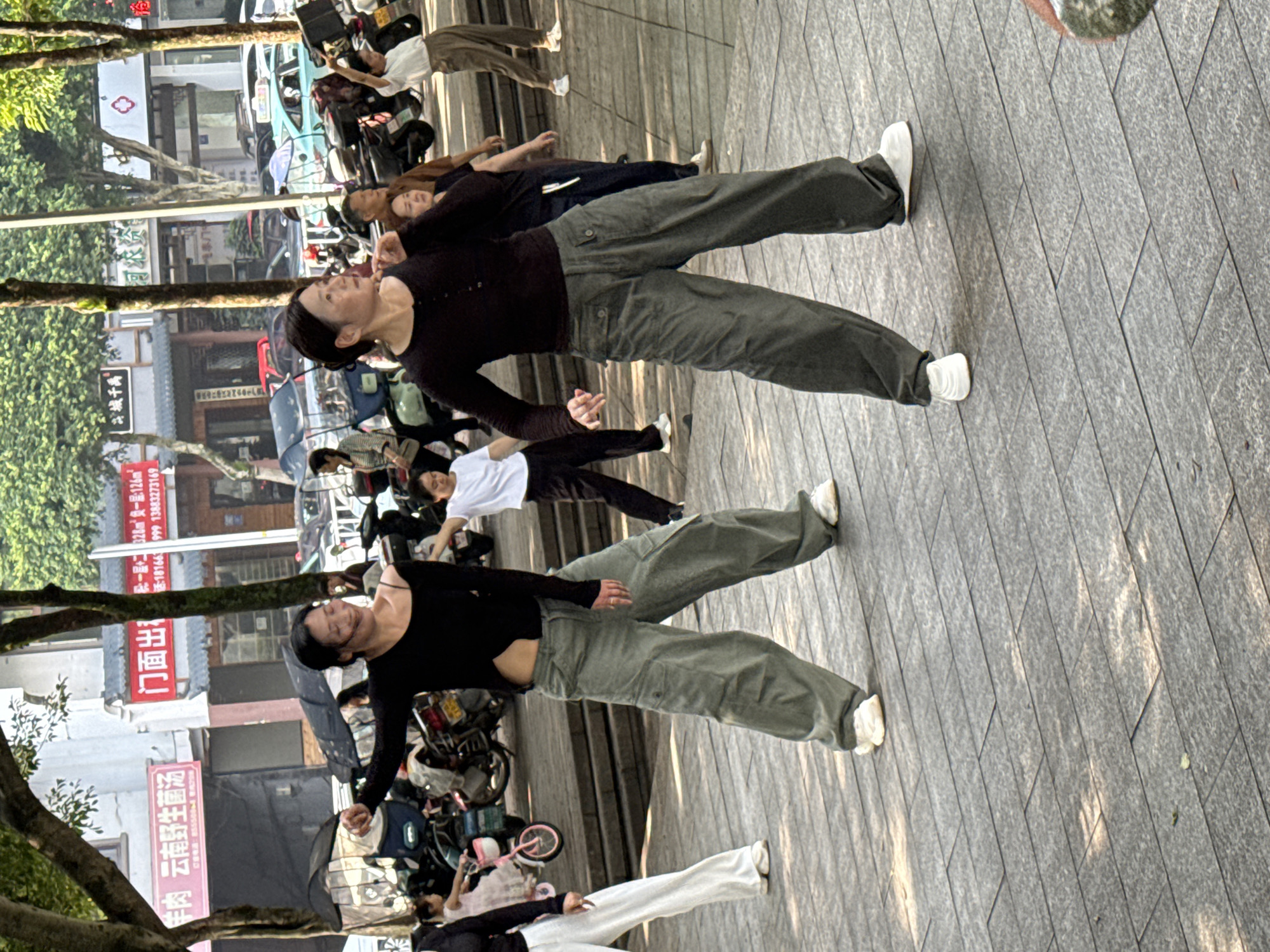 In Jiangjin, China, a group of women are practicing a dance routine outdoors in front of a row of buildings. Two women are prominently featured in the foreground, wearing loose-fitting pants and dark tops, with their arms extended as they move. Several other individuals are visible in the background, some participating in the dance and others observing. Various motorcycles and a bicycle are parked behind the dancers. The ground is paved with large, rectangular stone tiles, and the scene is illuminated by sunlight filtering through the trees overhead, casting dappled shadows on the pavement. The buildings in the background appear to be shops or businesses.