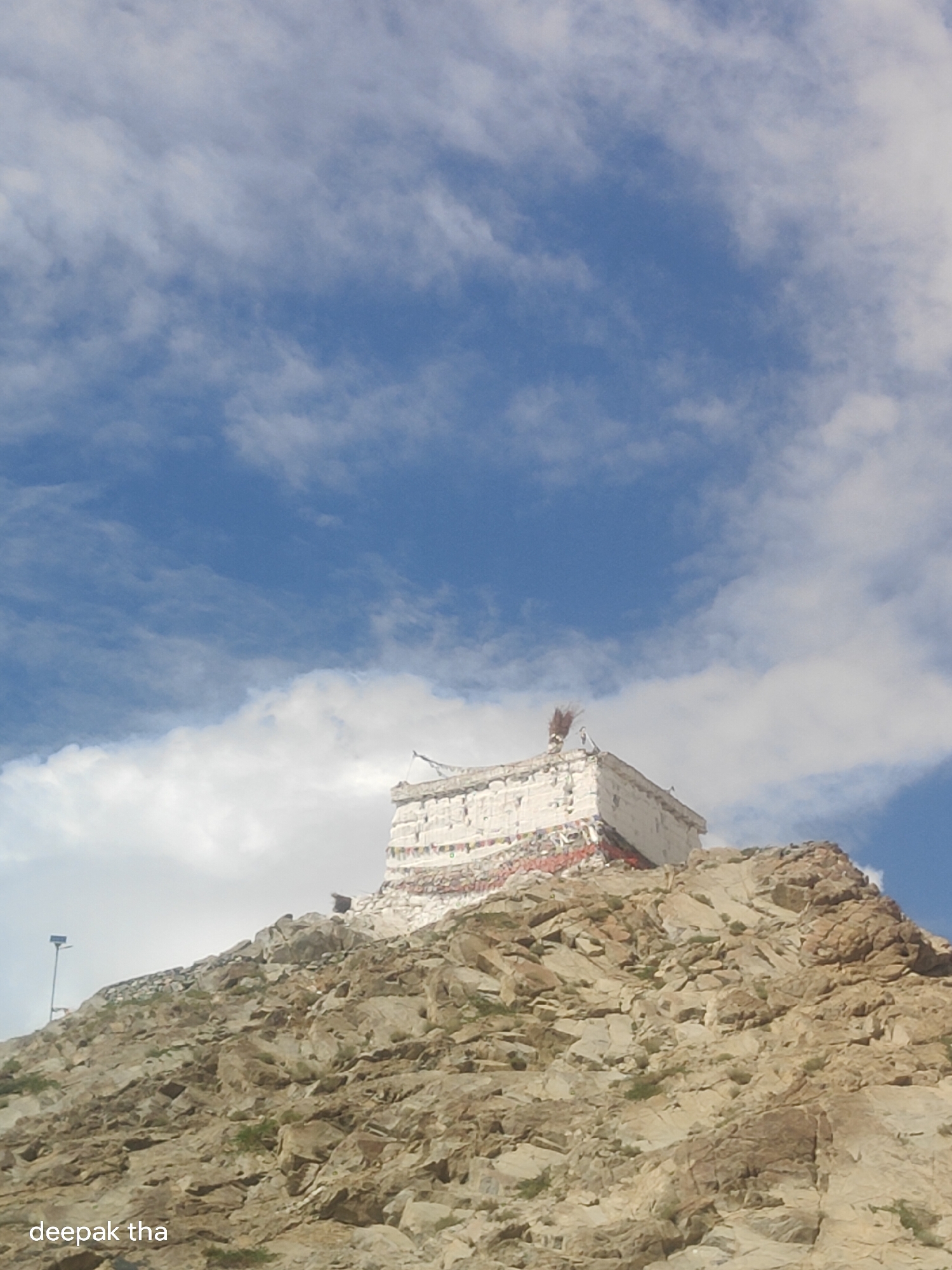 The image shows a view looking up at a white building perched on a rocky hill. The building has colorful prayer flags hanging from its sides and a broom-like object on its roof. The hill is covered in a rocky, uneven terrain with sparse vegetation. The sky above is blue with scattered white clouds. A lamppost is visible on the left side of the hill. The text "deepak tha" is superimposed on the bottom left corner of the image. This building is likely the Thiksey Monastery, located in the city of Leh, India, in the Ladakh region.