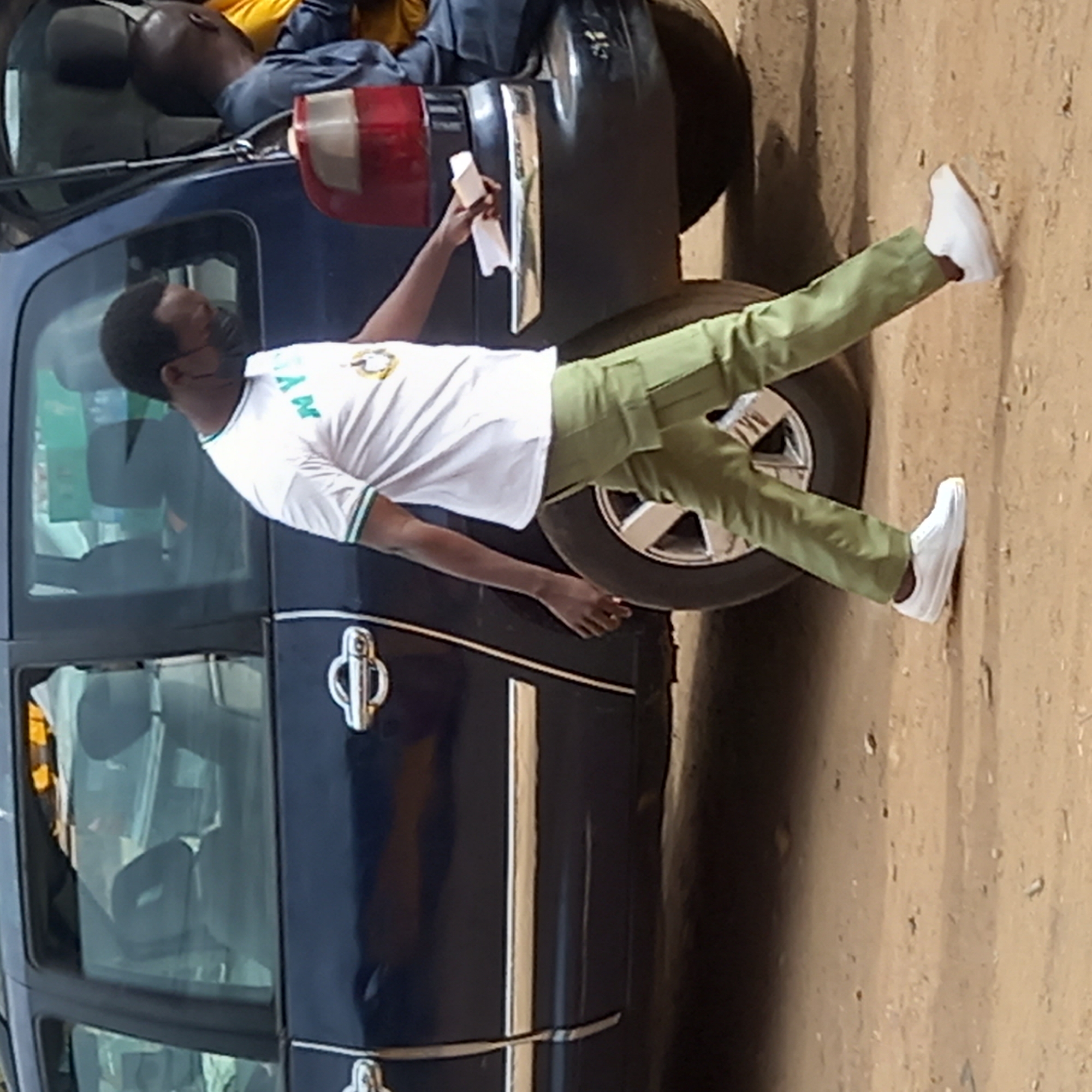 An outdoor, daytime image depicts a young man leaning against the side of a dark blue car on what appears to be a dusty or unpaved road. The man is wearing a white short-sleeved shirt with green and white trim, olive green trousers, and white sneakers. He has dark hair and is wearing a black face mask. On his shirt, the letters "NY" and a circular emblem are visible on the left chest area, suggesting a uniform, possibly related to a youth service corps. He holds a white rectangular object, possibly paper, in his right hand. His left arm rests on the car's body, and his right leg is extended with his foot on the ground near the car's tire, which has a silver hubcap.
In the background, another person is partially visible, appearing to be lying or slumped on the roof of the dark blue car, wearing a yellow shirt and dark trousers. The car has visible chrome door handles and a red taillight on its rear side. The scene suggests a casual environment in Dadi Riba, Nigeria. The lighting indicates a bright, clear day.