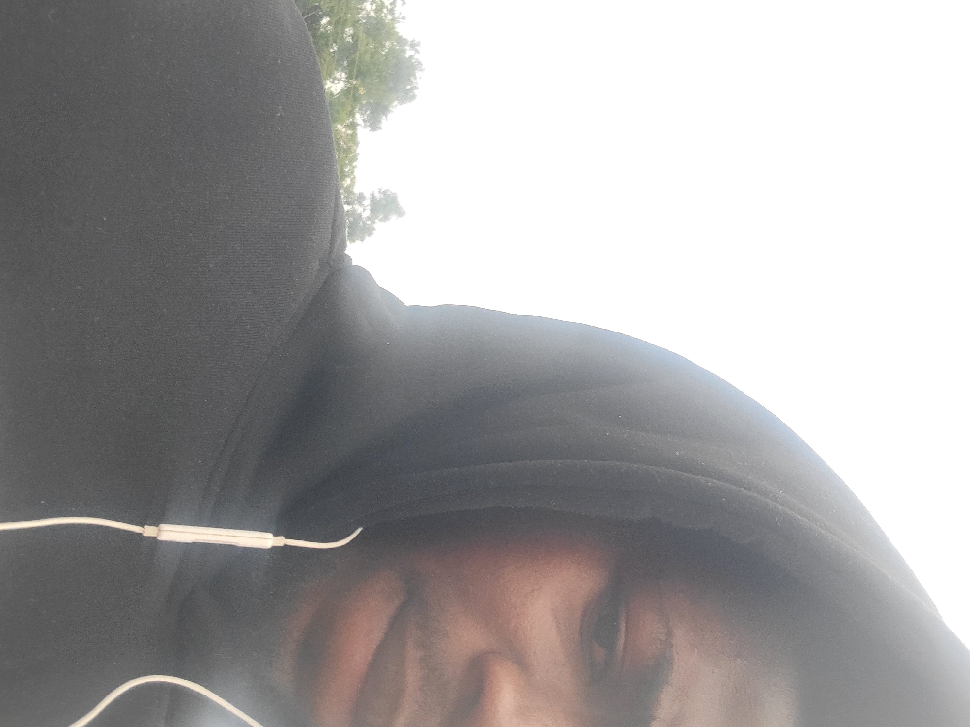 A close-up shot shows a person's face and shoulders, wearing a black hooded sweatshirt and white earbuds. The person's face is partially obscured by the hood and appears to be looking to the right. In the background, blurred trees and a bright, overcast sky are visible. The scene is outdoors, possibly in a park or wooded area, with no specific city or identifiable landmarks present.