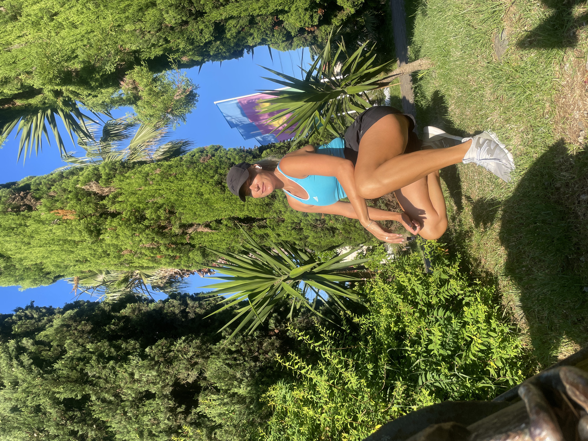A woman is crouching in a park setting, surrounded by lush green foliage and tall trees, with a clear blue sky overhead. She is wearing a turquoise tank top, black shorts, a black baseball cap, and gray athletic shoes. Her left arm is bent, with her hand resting on her knee, while her right arm is extended forward, with her hand resting on the ground. She is looking towards the right side of the frame with her eyes closed, and appears to be enjoying the sunlight. Two flags with colorful designs are visible in the background, behind a palm-like plant. The ground is covered in grass, and there are shadows cast by the trees and possibly other objects not fully visible. The overall impression is one of a serene and active moment outdoors. This scene is not in Sochi, Russia, but rather a tropical or Mediterranean climate.