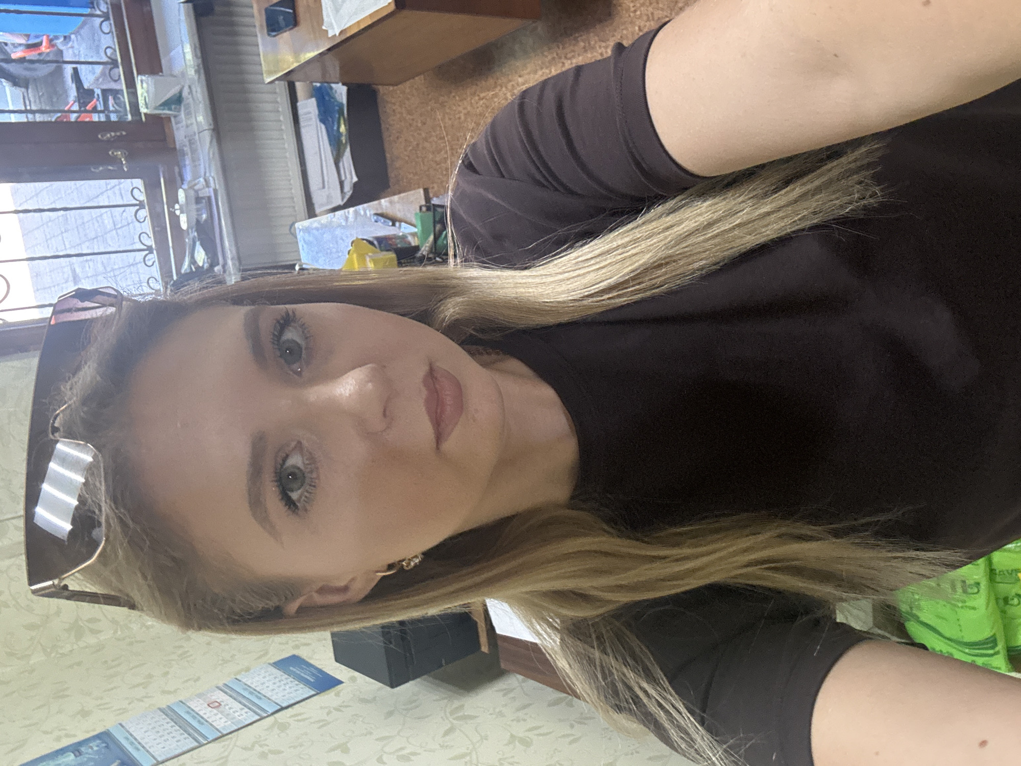 The image shows a close-up, selfie-style portrait of a young woman with blonde hair. She is wearing a dark-colored t-shirt and sunglasses are perched on top of her head. Her left eye is visible, and she is looking towards the camera. The background appears to be an indoor environment, possibly an office or room, with a calendar on the wall, a window with bars, a desk with various items, and a radiator. The lighting suggests it might be daytime. The location is described as Unknown, Unknown.