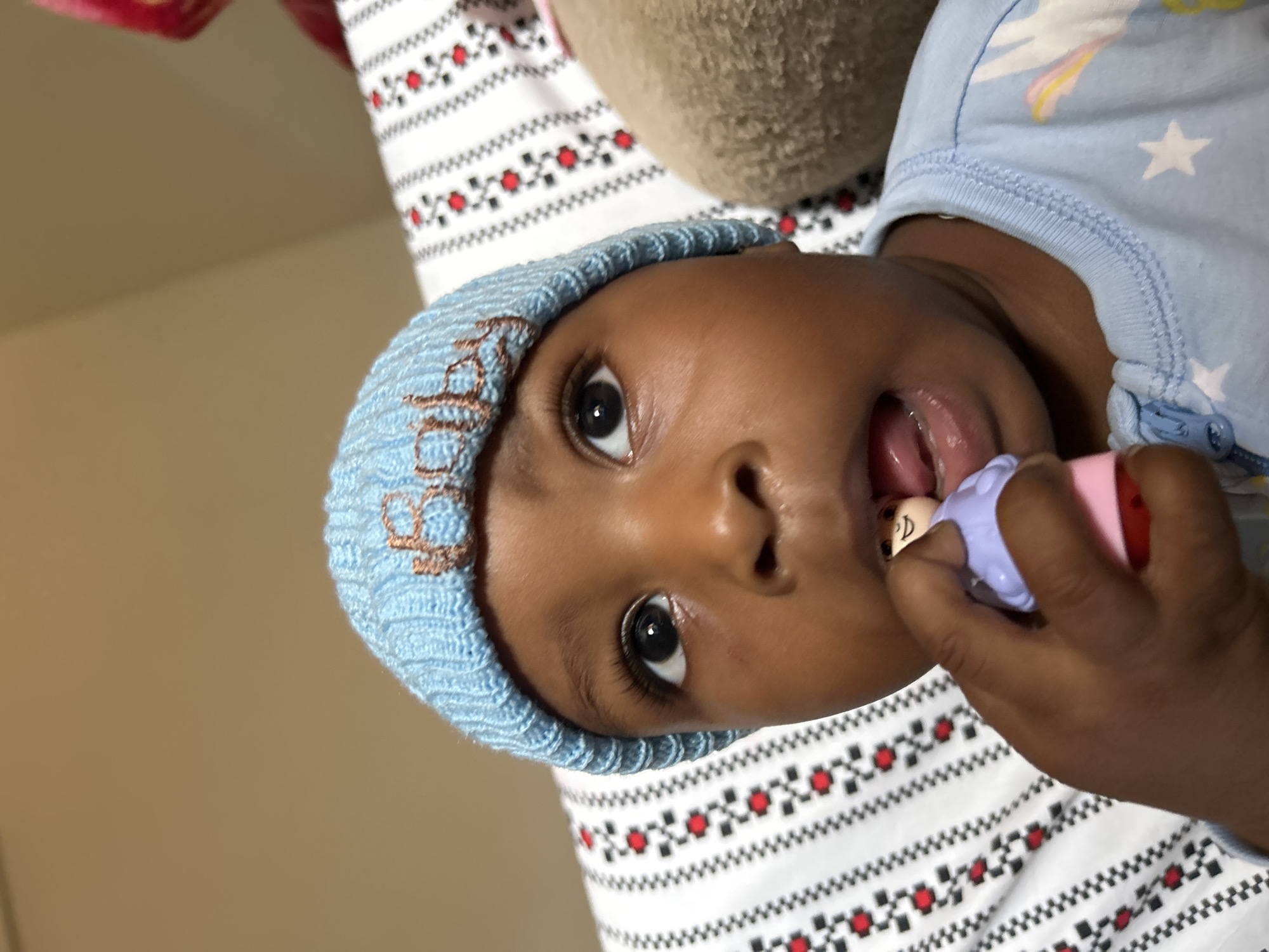 In this close-up, vertical shot, a dark-skinned baby is the central subject. The baby is wearing a light blue knitted beanie with embroidered brown lettering that appears to spell "Baby". They are also dressed in a light blue onesie adorned with white stars and whimsical unicorn-like figures, suggesting a playful and perhaps bedtime theme.
The baby's face is tilted, with one eye clearly visible, showcasing a dark pupil and iris. Their mouth is slightly open, and they are holding a small toy in their hand, which they are bringing towards their mouth. The toy is primarily purple and pink, with a small figure of a person on its end, resembling a pacifier or teething toy.
The background consists of a white fabric with a repeating pattern of black zig-zags and red circular shapes, possibly a blanket or bedding. A soft, beige object, perhaps a stuffed animal or pillow, is visible behind the baby's head. The overall lighting suggests an indoor setting, possibly a home environment. The scene appears calm and intimate, focusing on the baby's engagement with their toy. There are no immediate cues to suggest the time of day or weather conditions. The location context of Jos, Nigeria, is provided but not directly discernible from the visual elements of the image.