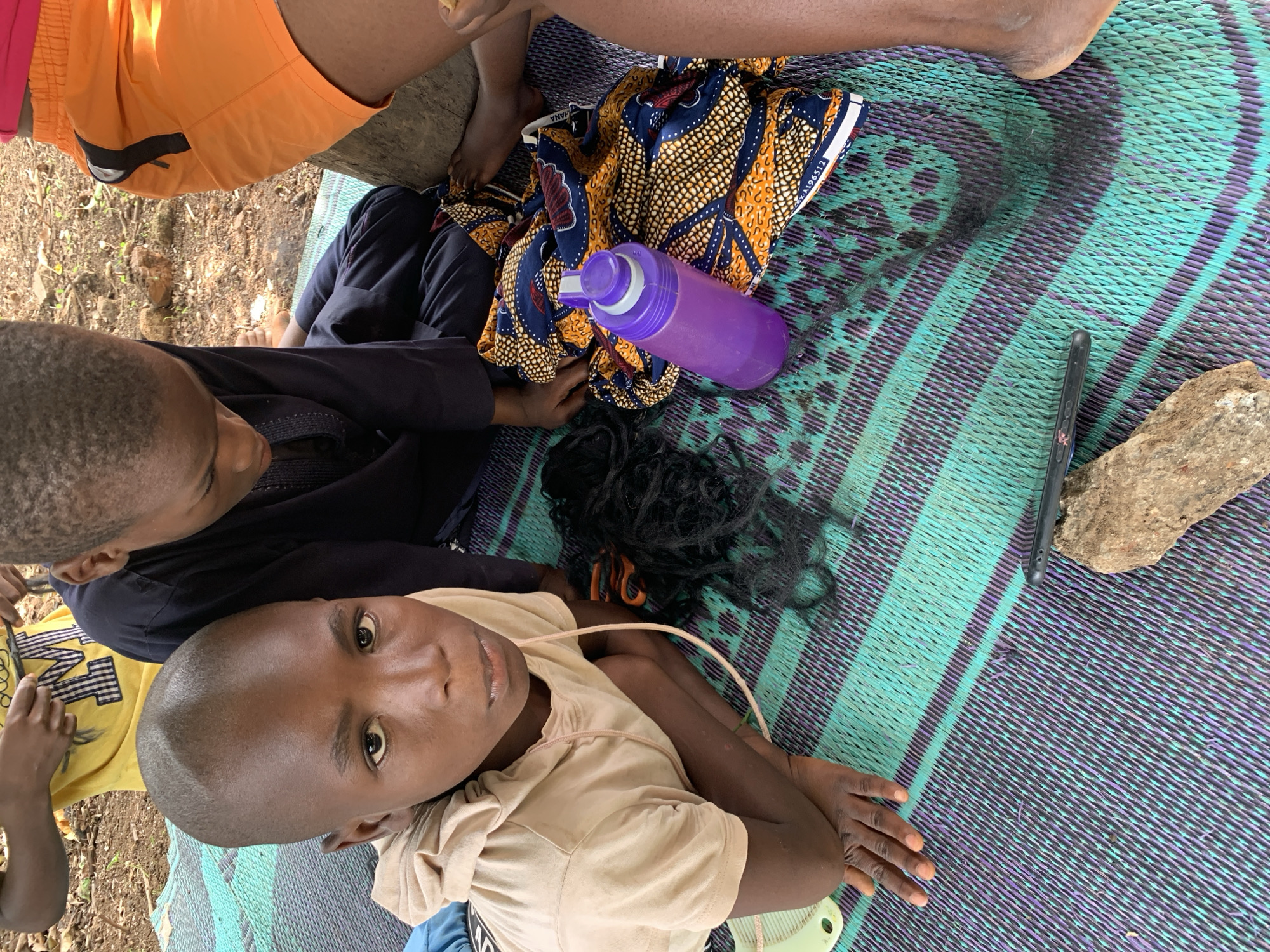 From an overhead perspective, the image shows a group of people gathered on a patterned mat in what appears to be an outdoor setting, likely in Akwanga, Nigeria. A child in a tan shirt looks up at the camera, while another person in a black shirt looks down. An individual wearing an orange shirt is partially visible in the upper left corner.
On the mat, there are several objects: a colorful patterned cloth, a purple water bottle, a dark wig, a cell phone, and a rock. It appears someone might be in the process of doing someone else's hair. The environment seems to be a yard or outdoor space, evident from the ground visible in the corners.