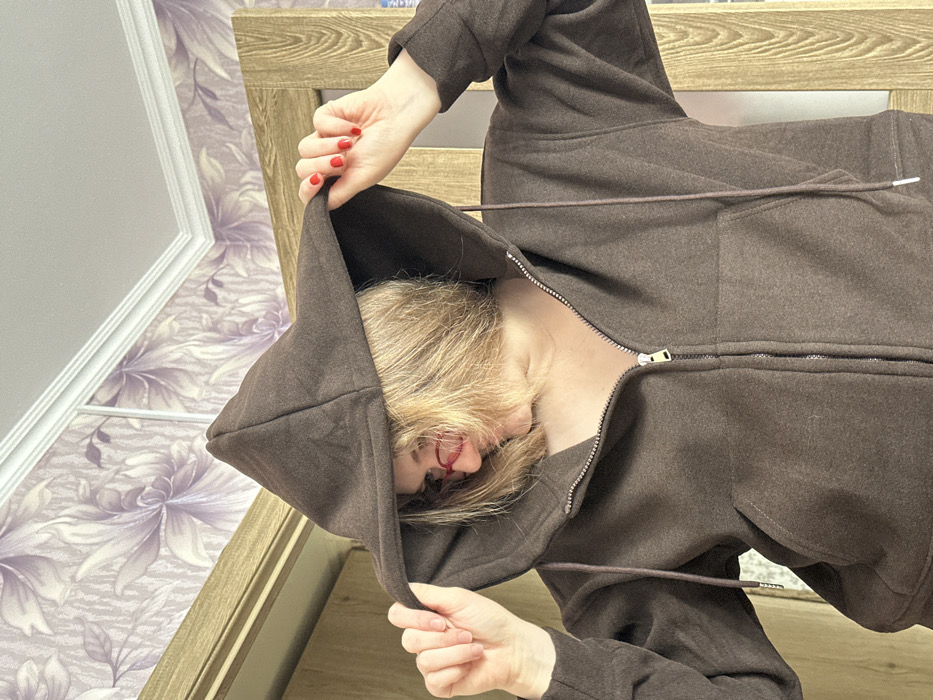 The image portrays a female individual in an indoor setting in Nizhnekamsk, Russia. The subject, viewed from the chest up, has shoulder-length blonde hair and wears red-framed glasses. She is dressed in a dark brown textile hooded zippered jacket, with the zipper partially open. Her hands, featuring red nail polish, grasp the brim of the hood, pulling it forward over her head. The background consists of a wall covered in light purple wallpaper with a large floral pattern, topped by white crown molding. Sections of two wooden door frames are visible; one light-colored to the left and a darker frame with a glass panel to the right.
