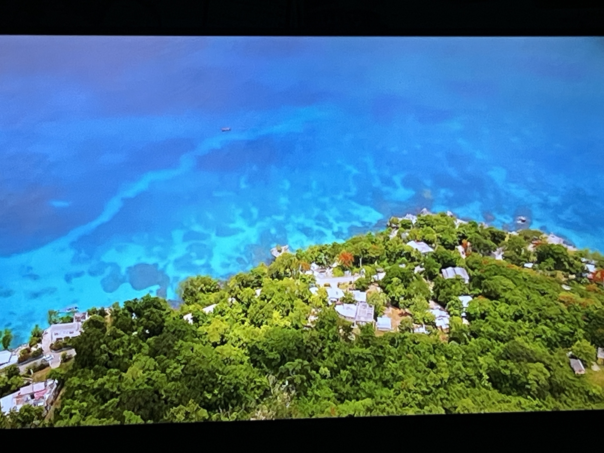 The image is an aerial view of a tropical coastline with lush green trees and buildings nestled amongst them. The ocean is a vibrant blue, with lighter patches indicating shallow water or coral reefs. There is a small boat visible in the water. The buildings appear to be residential or resort properties, with white roofs and some with visible landscaping. The scene evokes a sense of a secluded, picturesque location.
There is no information in the image that indicates it is located in Kharkiv, Ukraine. Kharkiv is a landlocked city in eastern Ukraine and does not have a coastline with tropical vegetation and clear blue ocean waters as depicted in the image.