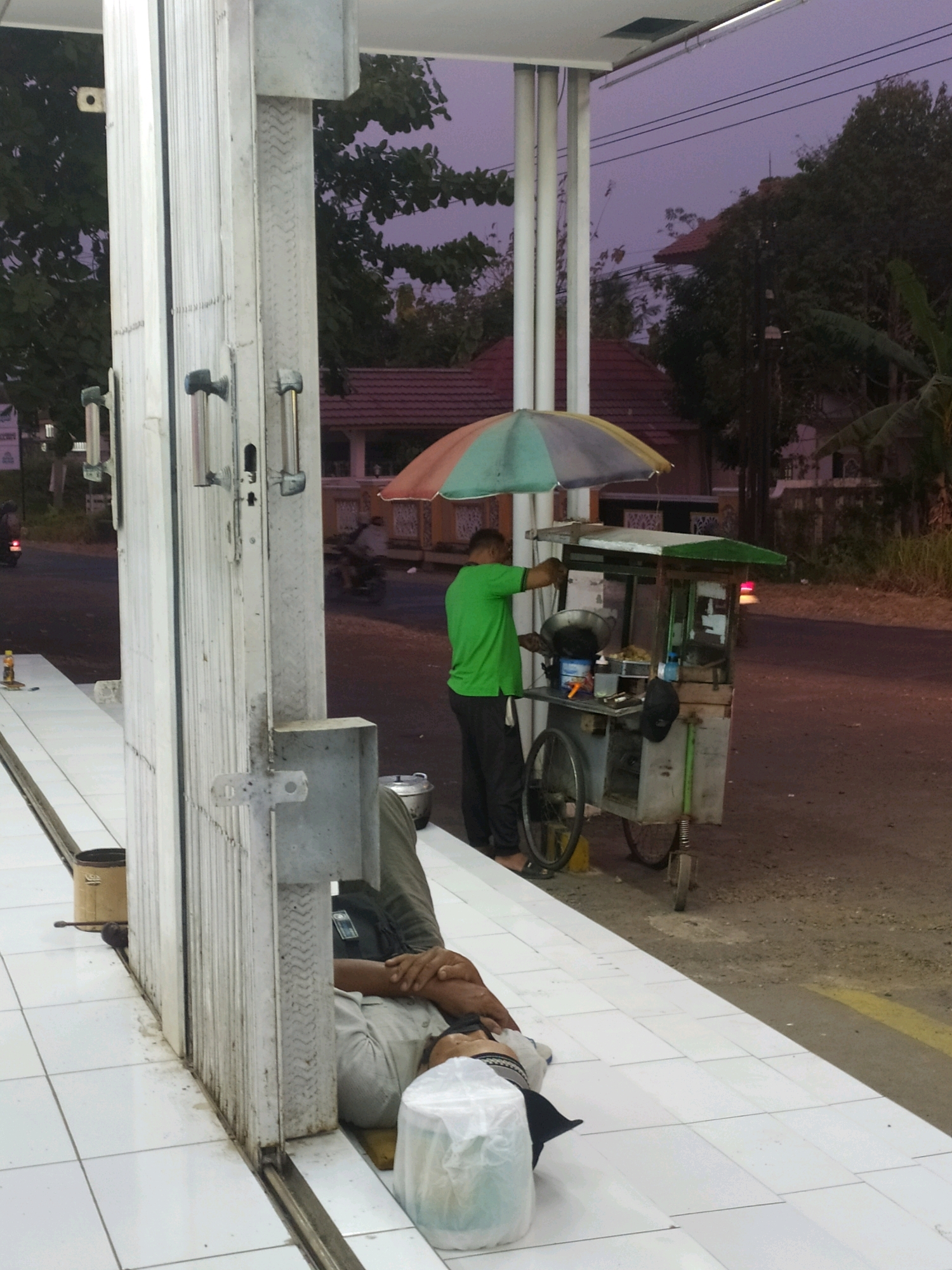 A man in a green shirt and black pants is operating a food cart at a gas station in Tambaksogra, Indonesia. The cart has a rainbow-colored umbrella and appears to be selling food. In the foreground, another man is lying on the tiled floor, seemingly asleep, with a white plastic bag and a yellow object near his head. The background shows trees, buildings, and power lines under a purple sky. The scene suggests a roadside food stall at dusk.