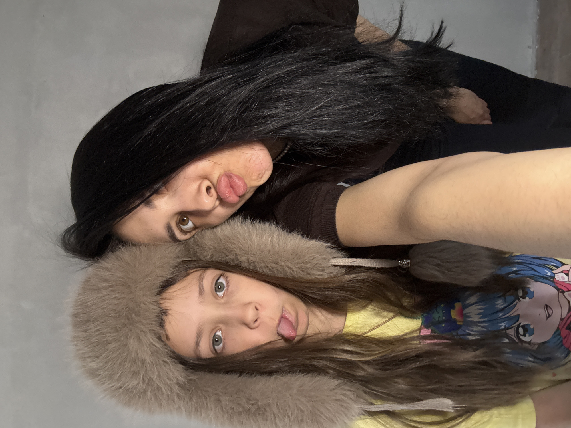 The image shows two females, likely an adult and a child or teenager, posing playfully for a close-up shot indoors. The photograph is oriented sideways, rotated 90 degrees clockwise.
The adult, positioned in the upper part of the frame, has long, straight black hair and is wearing a dark brown long-sleeved top. She has light brown eyes and is seen sticking her tongue out towards the camera in a humorous expression.
Below and slightly in front of her is the younger individual, who has long, wavy brown hair and is wearing a prominent light brown, fur-lined hood or hat. She is also sticking out her tongue, mirroring the playful gesture. Her eyes are light-colored, appearing to be green or blue-gray. She is wearing a yellow t-shirt with a colorful graphic of an anime-style character featuring blue hair and large eyes.
The background consists of a plain, light gray wall, suggesting an indoor setting. The overall mood of the image is casual, cheerful, and lighthearted, capturing a fun interaction between the two subjects.