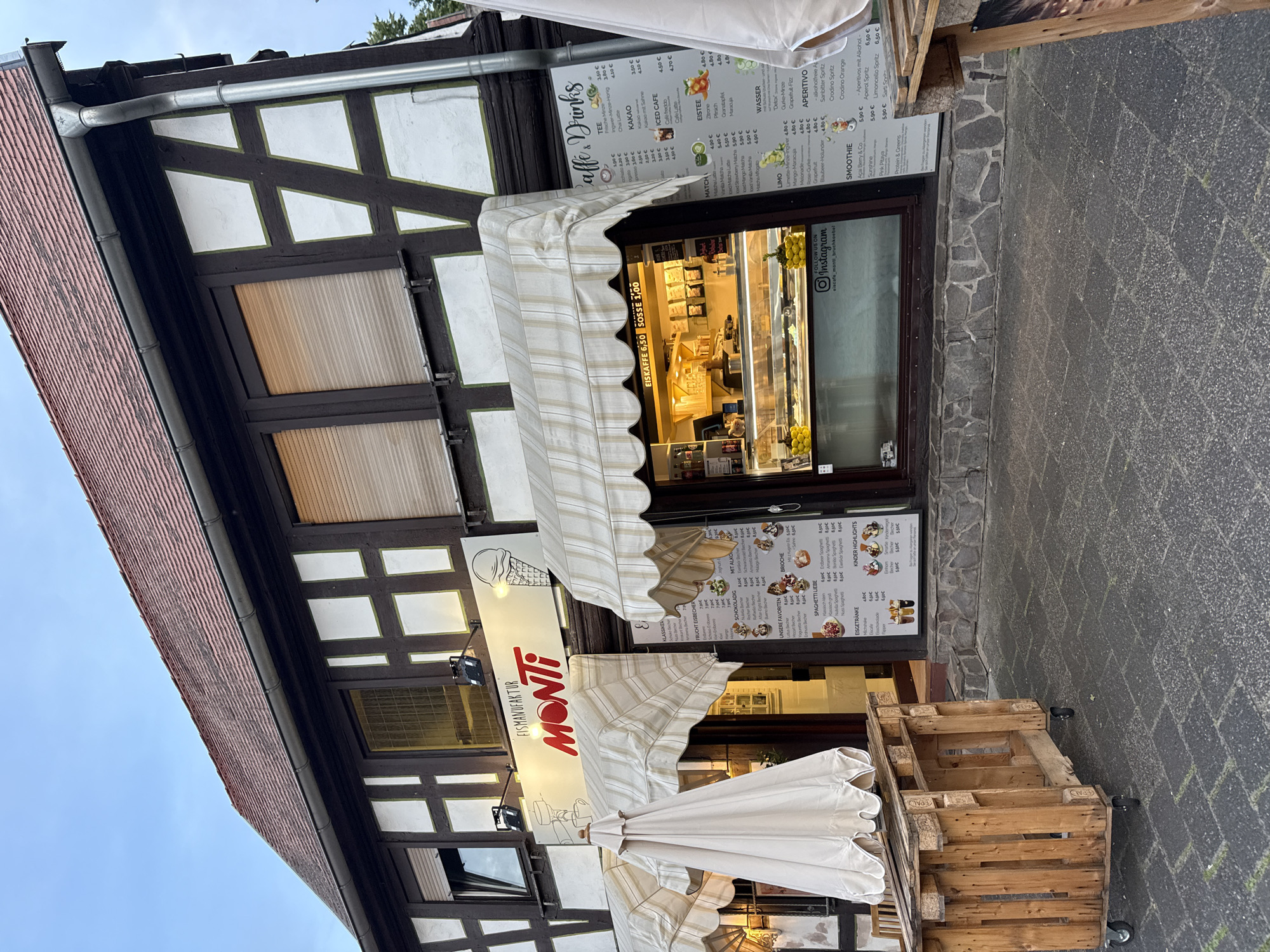 The image captures the exterior of "EISMANUFAKTUR MONTI," an ice cream shop located in Bruchköbel, Germany, on an overcast day. The building showcases traditional half-timbered architecture with dark brown beams contrasting with white and green-bordered infill panels. A reddish-brown tiled roof and a metal rain gutter are visible at the top.
The shopfront features a large display window and a glass door, revealing illuminated display cases inside, likely containing ice cream or pastries. Two white and beige striped awnings are extended above the window, providing shade, while another is folded. Large menu boards are prominently displayed next to the entrance, listing a variety of offerings in German.
Key menu categories include "Caffè & Drinks" (coffee and drinks), "TEE" (tea), "KAKAO" (cocoa), "ICED CAFE," "EISTEE" (iced tea), "MATCH," "WASSER" (water), "LIMO" (lemonade), "APERITIVO" (alcoholic and non-alcoholic aperitifs), "SMOOTHIE," and "Protein & Greens." Further boards detail "KLASSIKER" (classics), "FRUCHT EISBECHER" (fruit sundaes), "SCHOKOLADIG" (chocolate sundaes), "MIT ALKOHOL" (with alcohol), "UNSERE FAVORITEN" (our favorites), "BRIOCHE," "EISKAFFEE 6,50" (iced coffee for €6.50), "SOSSE 1,00" (sauce for €1.00), "SPAGHETTI LIEBE" (spaghetti ice cream), "KINDER HIGHLIGHTS" (children's highlights), and "EISGETRÄNKE" (ice cream drinks). Prices, denominated in Euros, are visible for many items, such as "Aperol Spritz 6,50 €" and "Erdbeer Spaghetti 8,50 €." A sign below the window reads "FOLLOW US ON Instagram @eiscafe_monti_bruchkoebel."
In front of the shop, the ground is paved with dark grey cobblestones, with a section of natural stone cladding along the building's base. Several wooden pallets, some with "EPAL" branding and equipped with wheels, are arranged near folded white outdoor umbrellas, suggesting outdoor seating or display. No people are visible, but the open shop and outdoor setup indicate business is active.