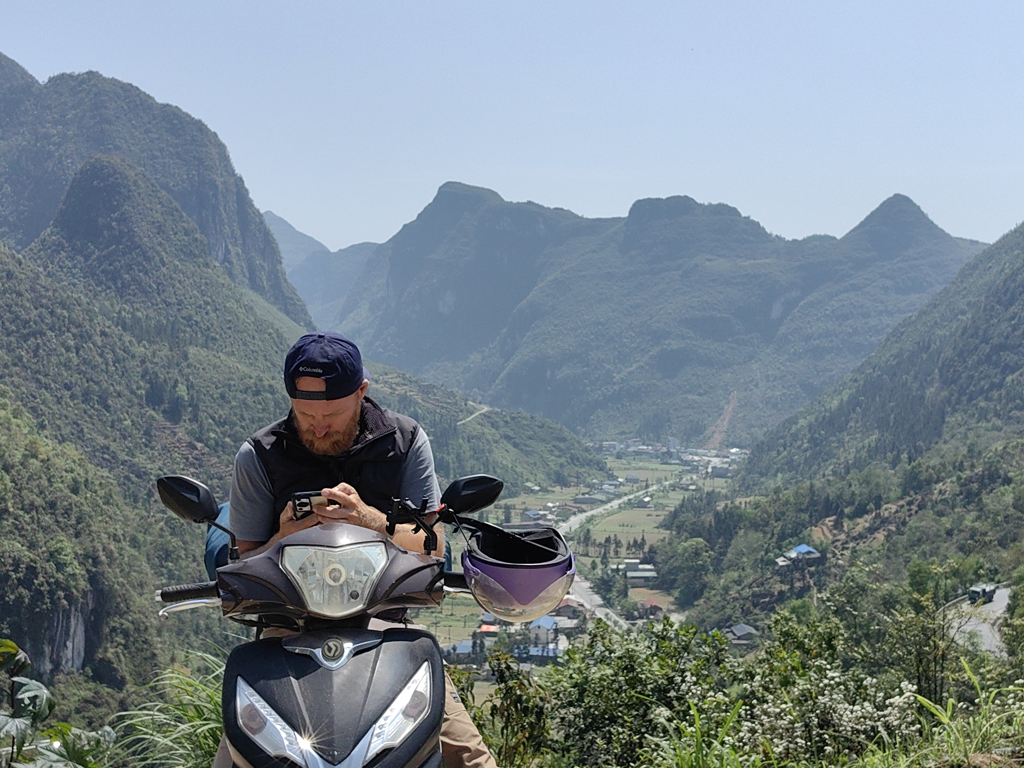 A man is sitting on a parked moped in front of a scenic mountain landscape in the Phố Bảng Commune, Vietnam. The man is fair-skinned, has a beard, and is wearing a blue baseball cap, gray shirt, and black vest. He is looking down at a phone in his hands. The moped is black and gray with a purple helmet hanging on the handlebar. In the background, rolling green mountains stretch into the distance, with a small town visible in the valley below. The sky is clear and blue. Vegetation is visible along the roadside.