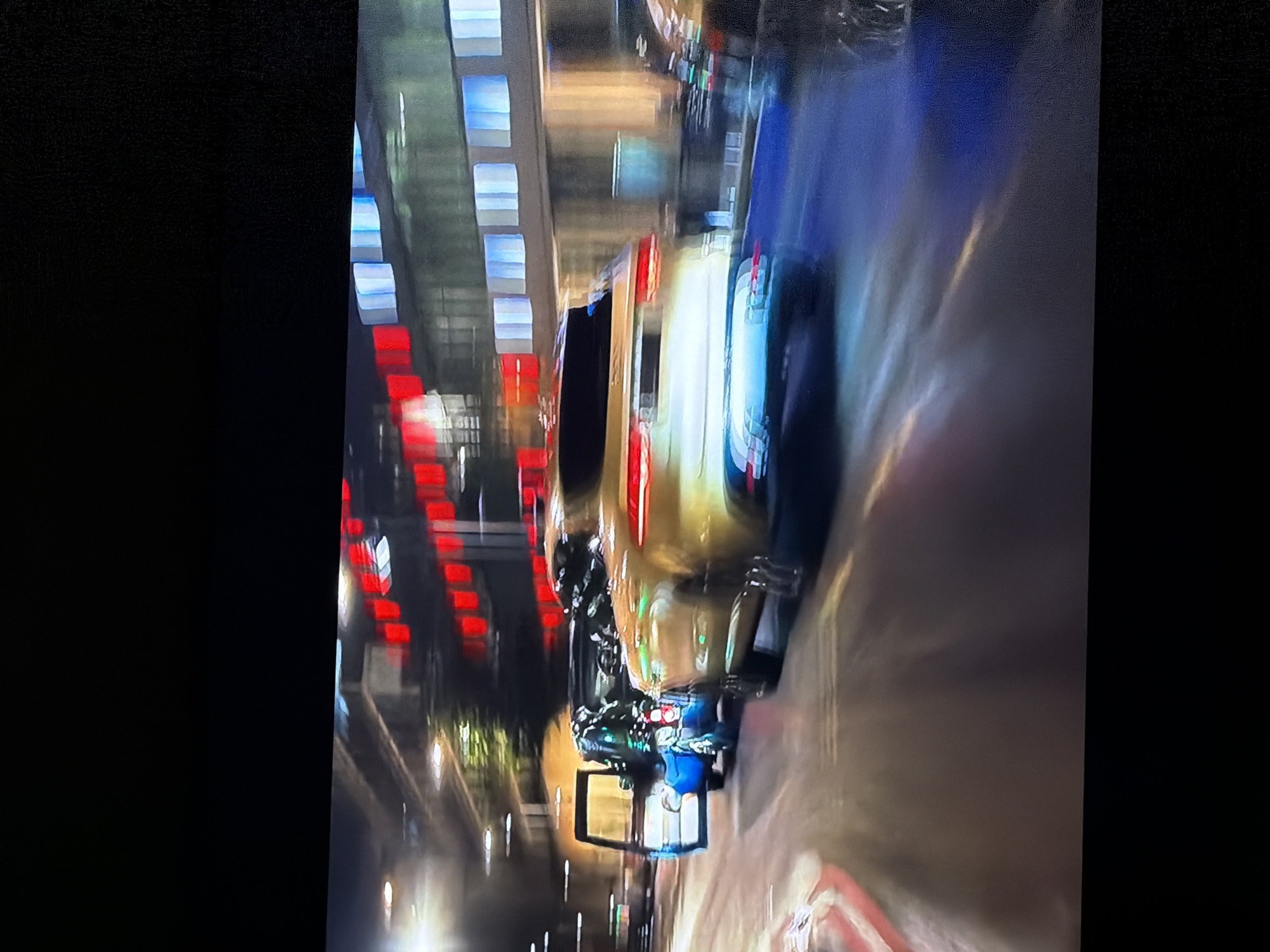 The image depicts a dynamic night street scene in Sofiivska Borshchahivka, Ukraine, characterized by significant motion blur, suggesting movement or a long exposure photograph. A golden or light-colored car, possibly a sedan or SUV, is prominently featured, seen from behind, and appears to be moving away from the viewer. Its rear lights glow red, creating streaky light trails across the blurred road surface. Several motorbikes or scooters are visible near the car, also exhibiting pronounced motion blur. In the background, multi-story buildings line the street, their numerous rectangular windows illuminated with cool blue and white lights, creating vertical streaks against the dark night sky. Other scattered lights, possibly streetlights or storefronts, add to the urban illumination. The scene conveys a sense of bustling city life at night. No individuals are clearly discernible, and no text is legible due to the motion blur. The overall atmosphere is energetic and illuminated.