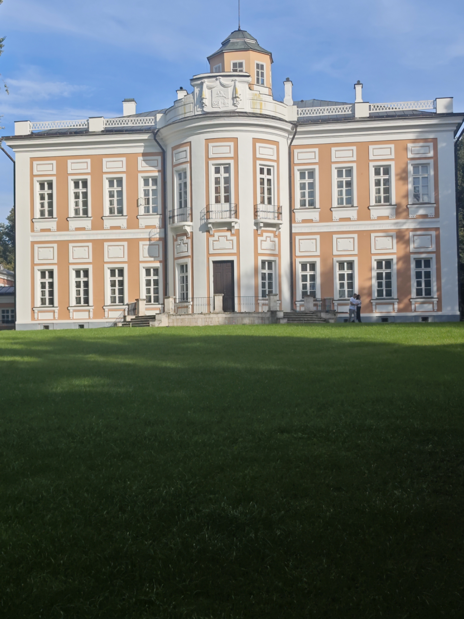 In Bolshiye Vyazyomy, Russia, a grand, two-story building with an orange facade and white accents stands on a manicured lawn. The building features a central rotunda topped with a cupola and ornate decorative elements. Numerous windows, many with white frames and panes, punctuate the facade. Two individuals are seen standing on the lawn near the building's entrance. The sky above is a clear blue with a few wispy clouds. The foreground is dominated by a lush green lawn, with shadows cast by unseen trees.