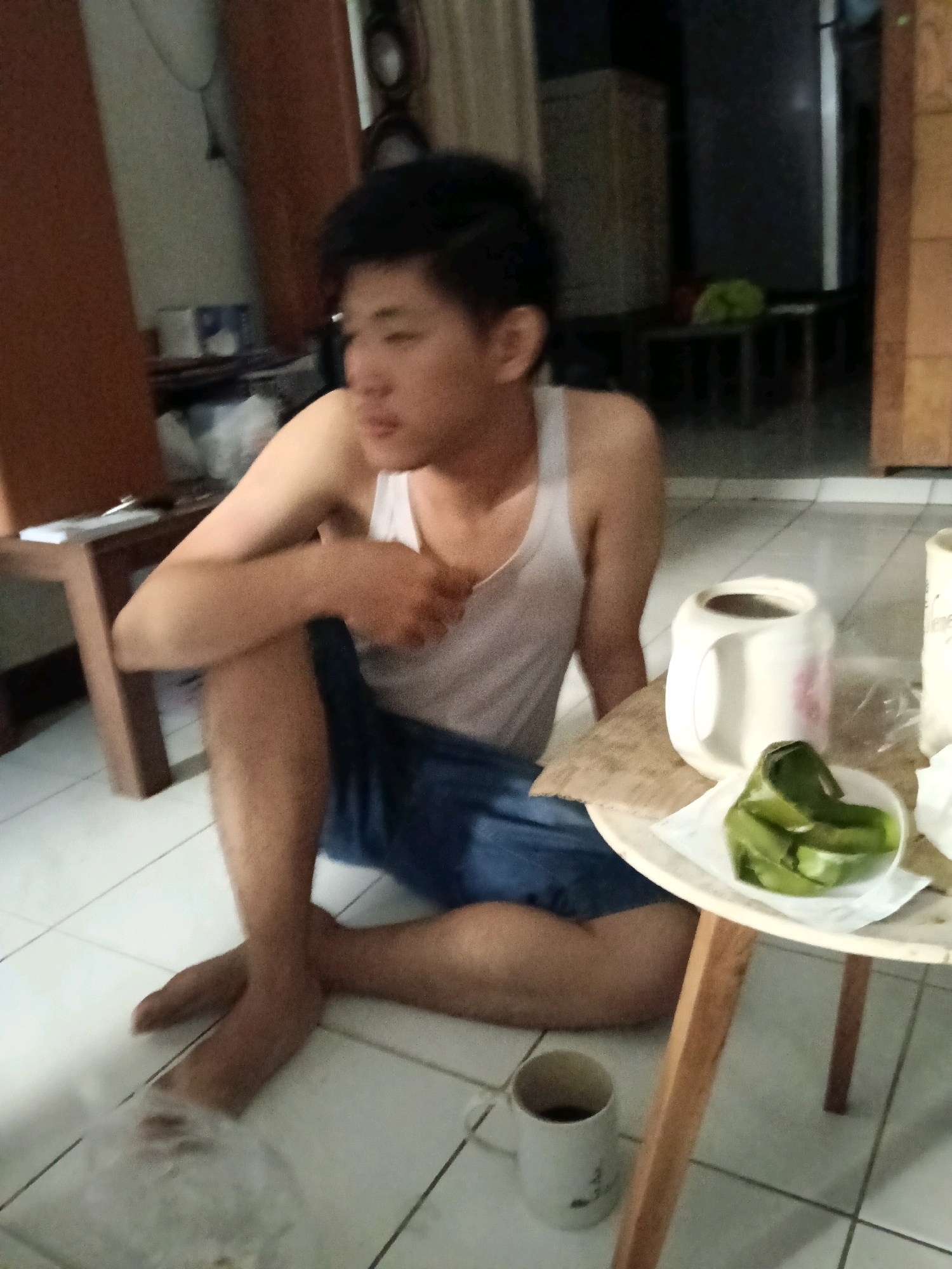 A young man with dark hair is sitting on a tiled floor, leaning against a wooden table. He is wearing a white tank top and blue shorts. His legs are crossed. In front of him on the table, there is a white mug with a pink design, a piece of brown paper, and a small white bowl containing several green items wrapped in a leaf. To the right of the mug, there's another white mug. The background shows a room with wooden furniture and a window with curtains. The lighting suggests it is daytime. The scene appears to be inside a residence in Jatimulya, Indonesia.