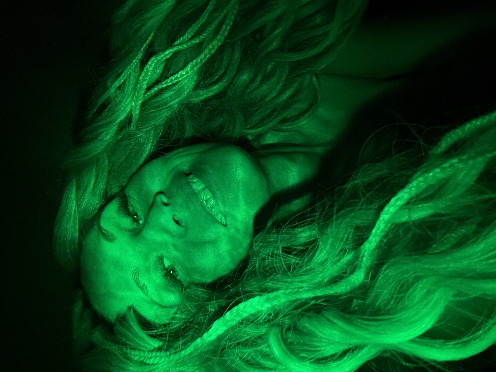 The image shows a woman lying down with her head tilted to the left. Her hair is long and braided, spread around her head. The scene is illuminated with a green light. The location is not discernable from the image.