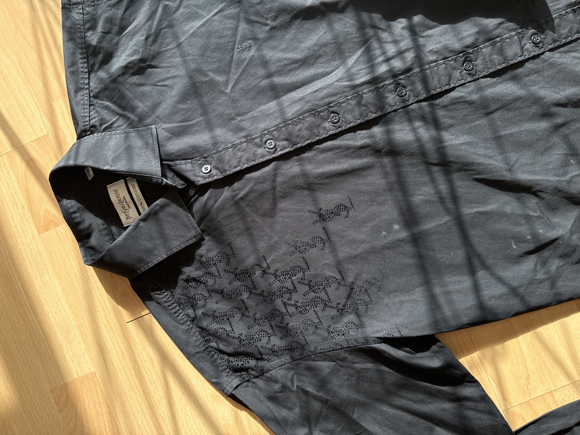 The image shows a black Yves Saint Laurent button-down shirt lying on a wooden floor. The shirt has a repeating pattern of the YSL logo made up of small dots on the front of the shirt. The shirt is wrinkled and has a tag on the collar that reads "Yves Saint Laurent" and "100% Cotton". Sunlight shines across the floor and the shirt, creating shadows. There is no indication of the location being London, United Kingdom.