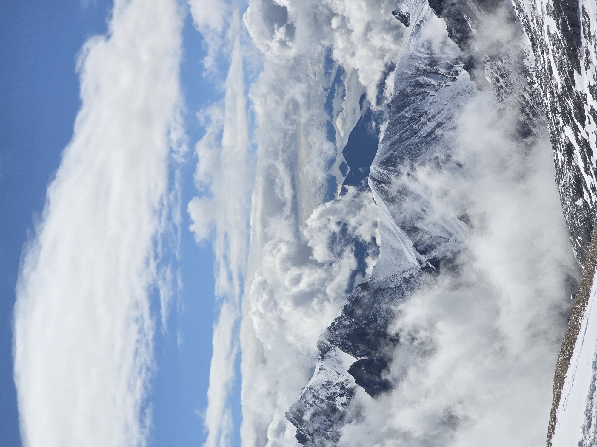 The image displays a high-altitude mountain landscape. Snow-covered peaks dominate the scene, with rugged terrain and sharp ridges. Clouds are prevalent, with large, white formations obscuring parts of the mountains and the sky. Patches of blue sky are visible through breaks in the clouds. The lighting suggests it is daytime, possibly mid-morning or afternoon, with the sun casting some shadows on the snow and rock. The foreground shows a rocky, snow-dusted slope. The background reveals layers of distant mountains, some of which are partially veiled by fog or clouds. There are no people or man-made structures visible in the image. The overall impression is one of a vast, wild, and imposing natural environment. This scene is not located in the city of Garmisch-Partenkirchen, Germany, but rather depicts a remote, high-mountain region.