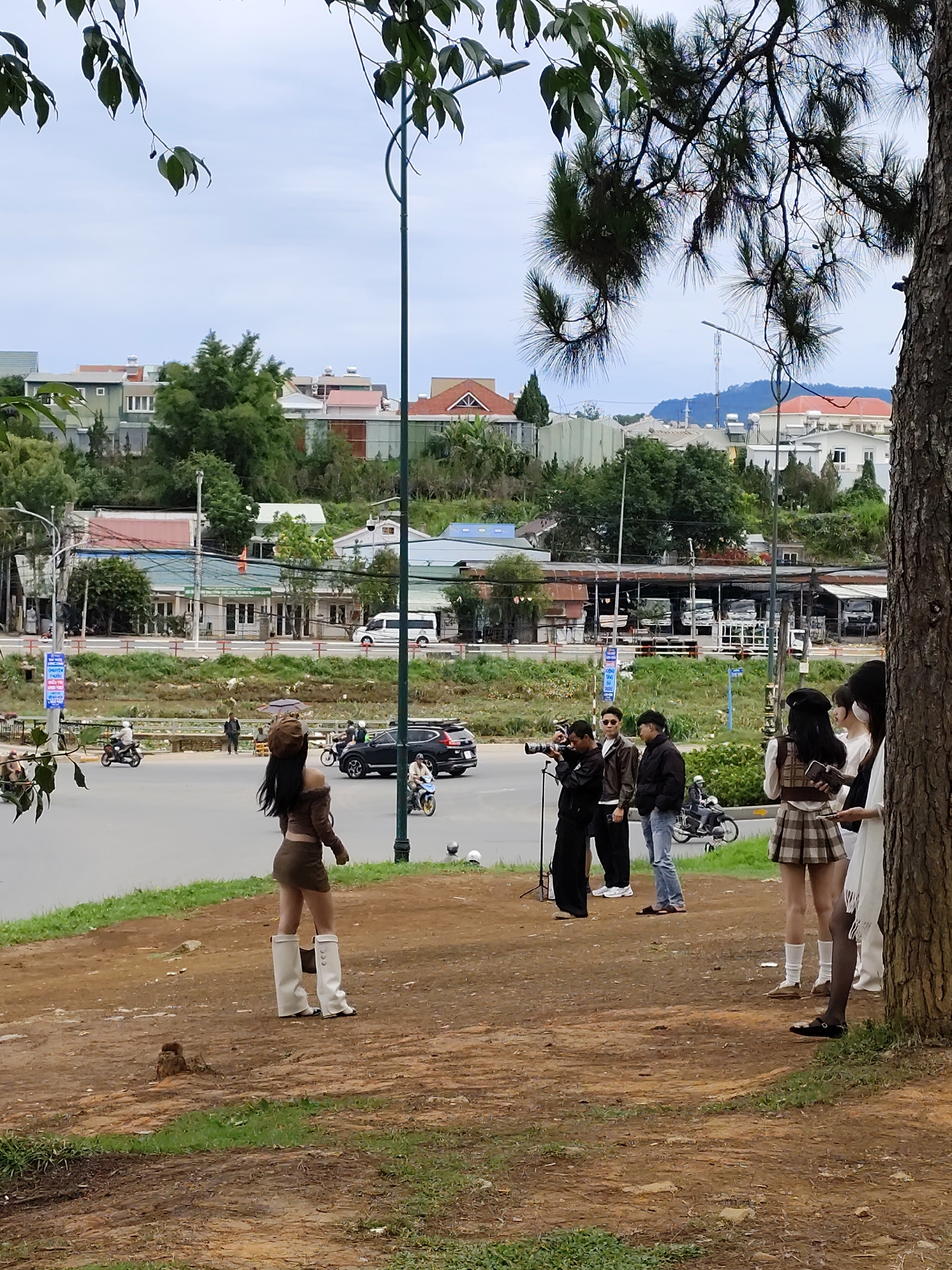 The image captures a scene in Da Lat, Vietnam. A woman in a brown outfit and white boots poses on a dirt patch near a road. A group of people, including a photographer with a camera on a tripod, are near her. Additional women stand closer to a tree on the right side of the frame. The road features light traffic with cars and motorcycles. Beyond the road, there is a low green area and then buildings, including houses and shops, that give context to the urban setting. The sky is overcast.
