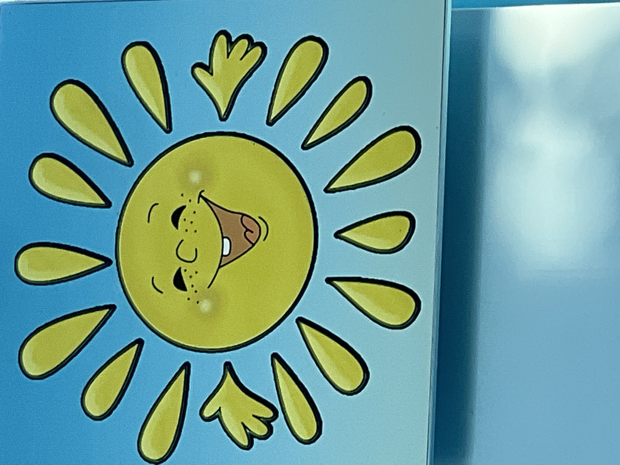 The image depicts a cartoon drawing of a sun with a smiling face, freckles, and rosy cheeks. It has bright yellow rays extending outwards and two small, yellow hands. The sun is positioned against a bright blue background. The bottom portion of the image shows a lighter blue surface, possibly a wall or table, with some reflection. The drawing appears to be part of a decoration, possibly in a child's room or educational setting. The image is not taken in Bender, Moldova.