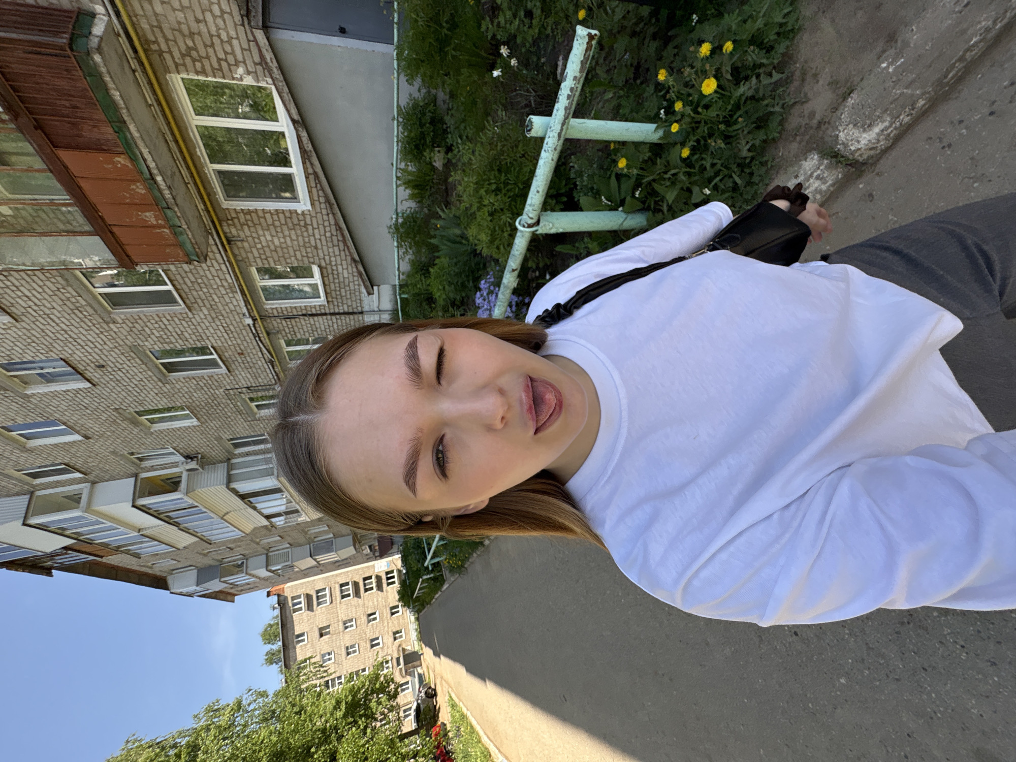 A young woman is depicted in a selfie outdoors, appearing to be in an urban setting. The background reveals a multi-story brick building and a paved sidewalk. The photo appears to be taken in daylight. Vegetation, including grass and some flowers, is visible. The woman is wearing a white, long-sleeved shirt. The woman sticks her tongue out. Based on the presence of buildings and Google Maps data, the location is identified as Chaikovsky, Russia.
