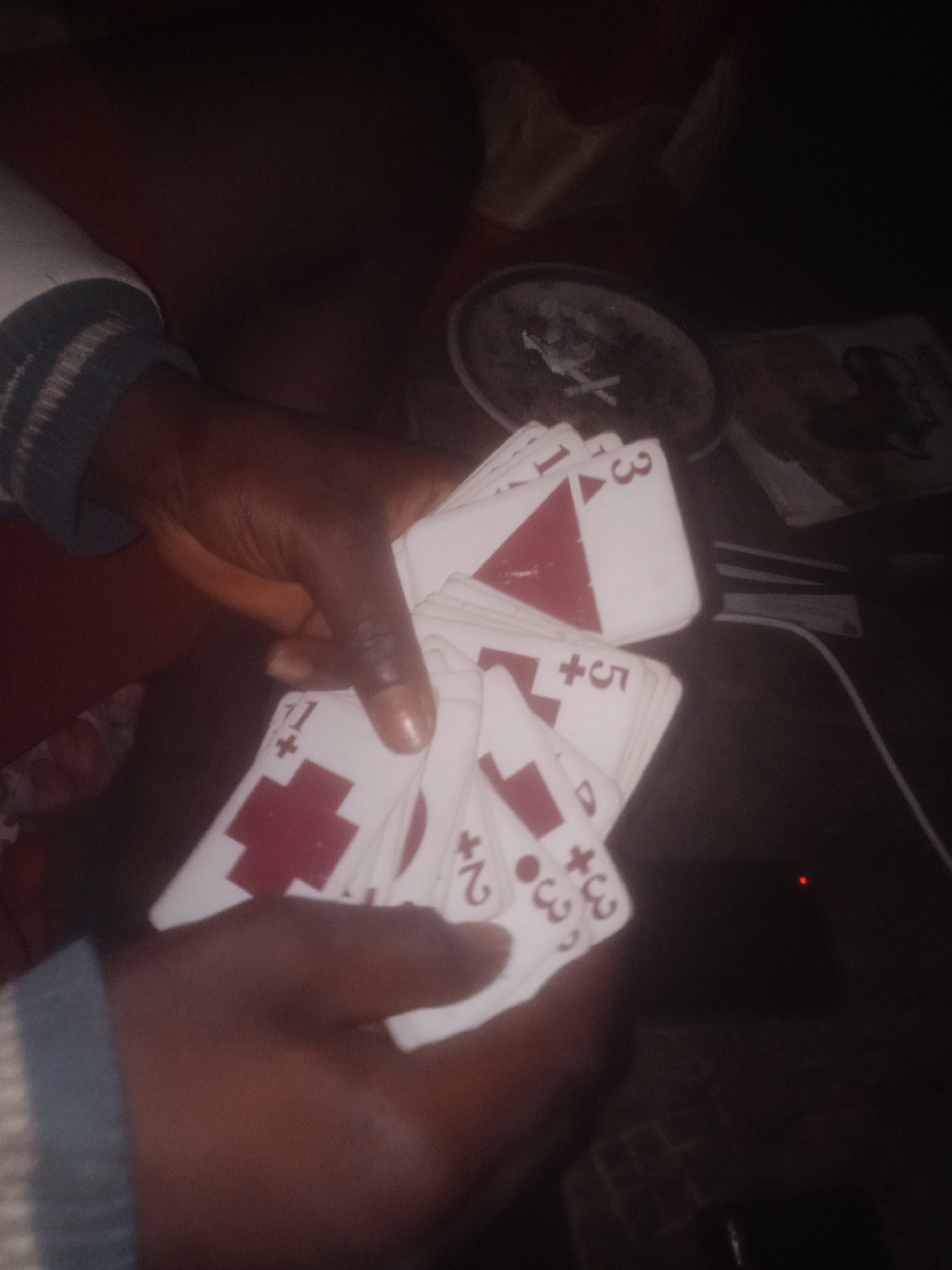 A dark, dimly lit image shows a pair of hands holding a fan of playing cards. The cards are white with red symbols, appearing to be some type of custom or non-standard deck. Some cards have large red crosses, while others have numbers and what look like triangles or abstract shapes in red. The hands are dark-skinned. The background is indistinct and dark, suggesting an indoor environment with low lighting. The overall impression is of someone in the process of playing or sorting through a deck of cards.