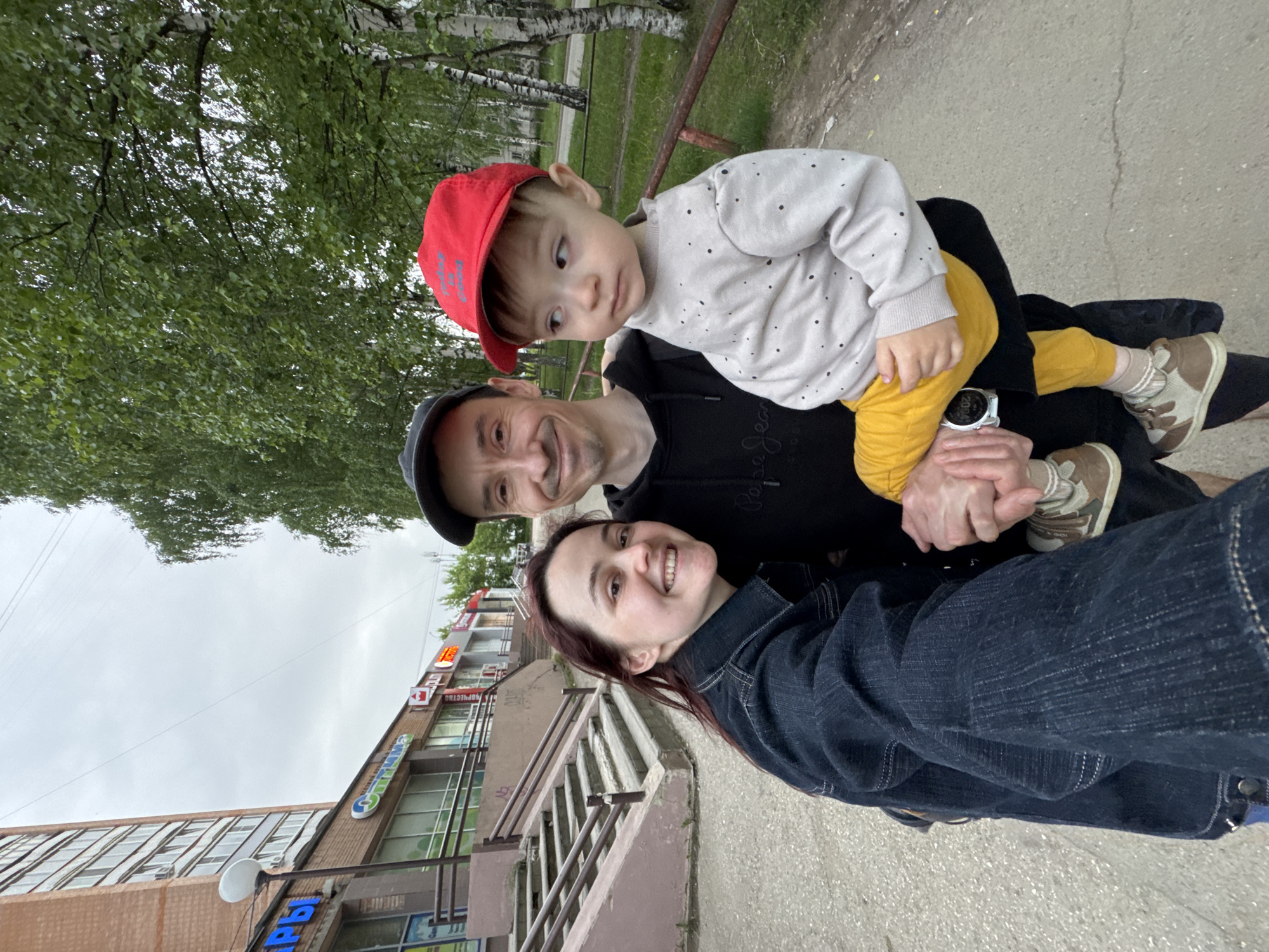 This image captures three individuals in an urban outdoor setting at Molodezhnaya Street, 18, Izhevsk, Udmurt Republic, Russia. The woman on the left, with dark reddish-brown hair, smiles directly into the camera while taking a selfie. She wears a dark blue denim jacket. Adjacent to her, a man with short dark hair, wearing a black baseball cap and a black hoodie with a "Pepe Jeans" logo, holds a small child. A silver-colored smartwatch on his left wrist displays "20:04". He exhibits a faint smile. The child, a toddler, wears a red baseball cap, a light grey long-sleeve top with small dark polka dots, bright yellow pants, and brown and beige sneakers; the child's gaze is directed towards the right side of the frame.
The background on the left features a multi-story brick building with commercial establishments on the ground floor, accessible by concrete steps with metal railings. Visible shop signs include "КАНЦТОВАРЫ" (Stationery), "Оптика" (Optics), "Красное и Белое" (Red & White, a retail chain), and "Магнит Косметик" (Magnit Cosmetic). A street lamp post is also visible. To the right, numerous green deciduous trees, consistent with birch trees, line an asphalt path, with a low wooden fence or railing separating the path from a grassy area. The sky is overcast, providing diffused lighting to the scene.