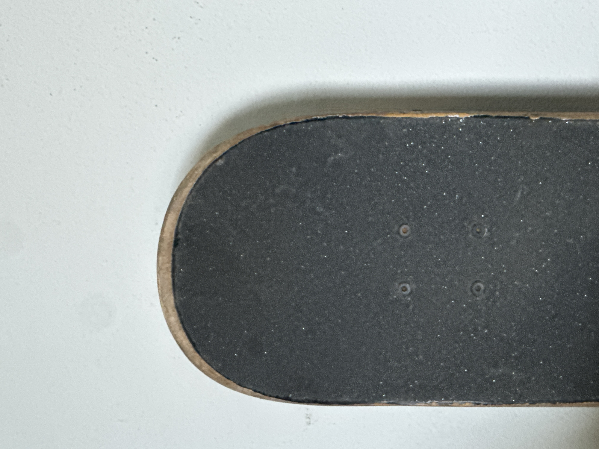 The image shows a close-up view of the tail end of a skateboard resting against a textured, light-colored wall. The skateboard is oriented vertically, with its rounded tail at the top of the frame. The grip tape, which is dark gray and speckled with white flecks, covers the surface of the tail. The edge of the skateboard deck, made of wood, is visible along the left and right sides. Four circular indentations, likely for truck bolts, are present on the grip tape. The wall behind the skateboard appears to be painted or textured plaster, with some subtle shadows and variations in tone. The overall lighting is soft, suggesting an indoor or shaded outdoor environment. There is no indication of the city of Karon, Thailand, in the image.