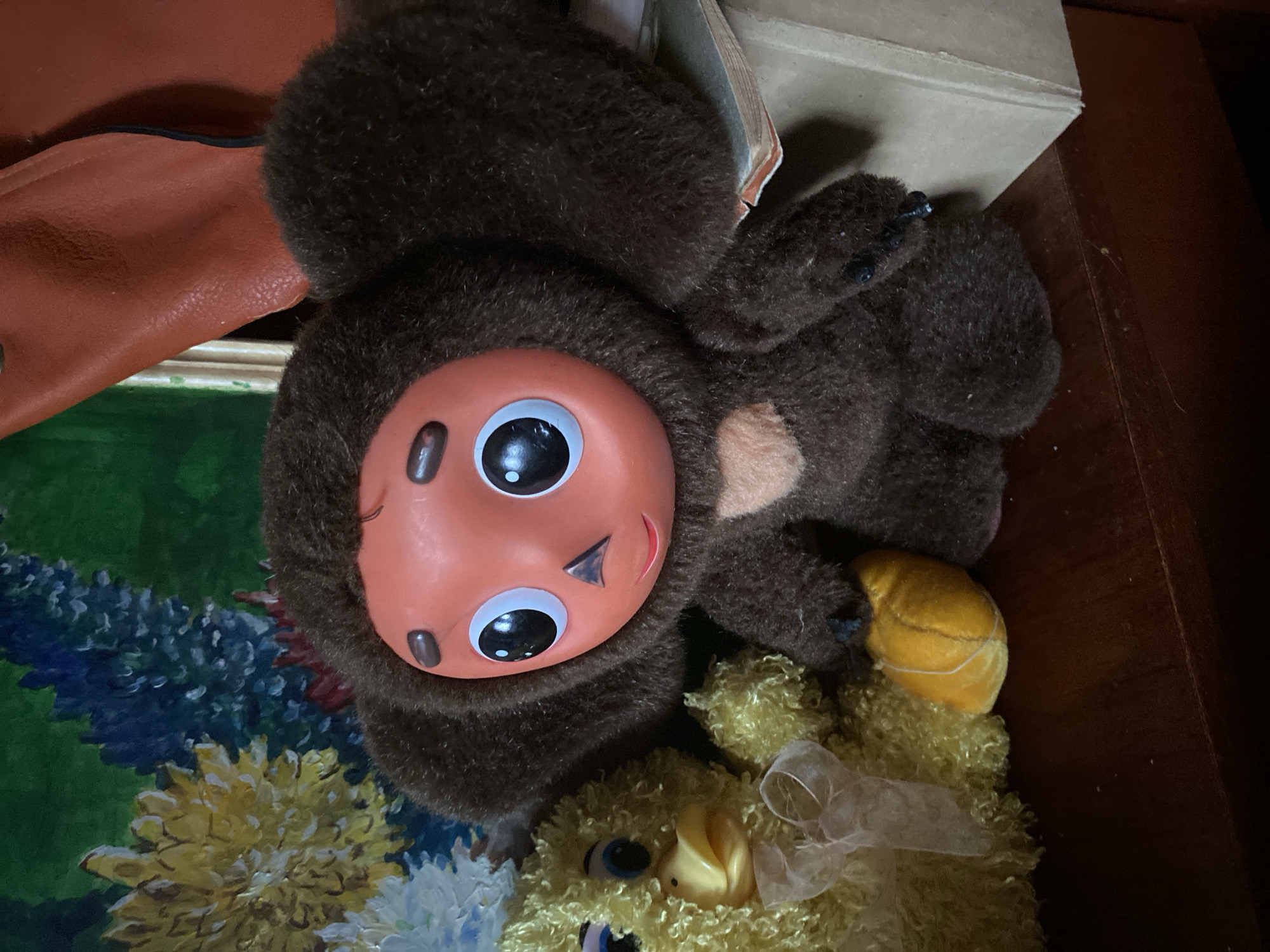 The image shows two stuffed toys on a wooden surface. On the left is a yellow, fluffy toy duck with blue eyes and a pale pink ribbon tied around its neck. To the right of the duck is a brown, furry toy with large, round ears and an orange face. It has large black eyes with white pupils, a small black nose, and a faint smile. This toy is holding a small yellow object in its right hand. In the background, there is a colorful painting of flowers and a brown leather object. The scene is indoors, likely a domestic setting. There are no people or specific identifying landmarks of Votkinsk, Russia visible in the image.