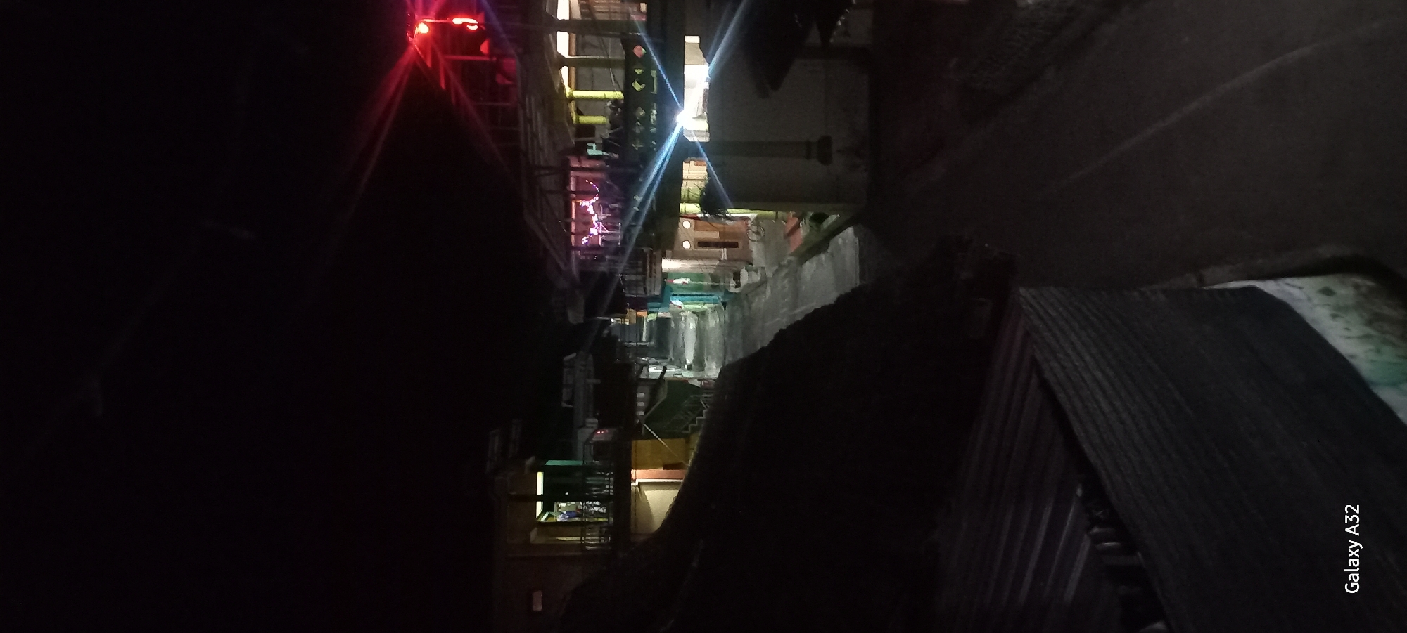 The image captures a street scene at night in Plumpung, Indonesia. The perspective is from an elevated viewpoint, looking down a narrow street lined with buildings. The sky is completely dark, indicating nighttime. Streetlights and lights from within the buildings illuminate the scene, casting long shadows.
On the left side of the street, there are multi-story buildings with visible windows and balconies. One building has an open balcony area with people inside, illuminated by interior lights. The architecture appears to be functional and residential or commercial.
On the right side of the street, there are also buildings, some with covered porches or balconies. Some structures have colorful facades, suggesting shops or small businesses. There are visible signs of lighting, including what appears to be a red light at the top of a pole on the far right. The street surface is paved and appears wet in places, reflecting the lights.
In the foreground on the left, there is a dark, sloped roof structure, possibly part of a dwelling or an adjacent building, which takes up a significant portion of the lower left quadrant of the image. The lower right corner of the image displays the text "Galaxy A32", indicating the device used to capture the photo. No people are clearly identifiable as distinct individuals, but figures can be seen inside some illuminated buildings, suggesting human presence. The overall impression is a quiet, residential or mixed-use street at night, lit by artificial lights.