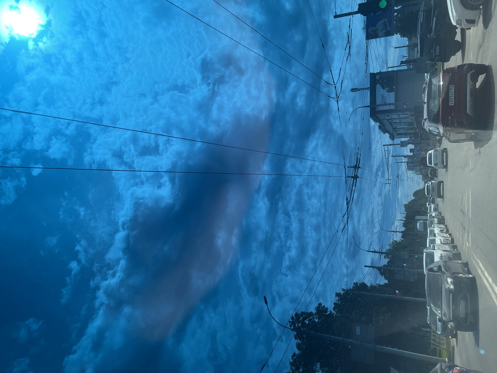 This media file, oriented vertically, captures an urban street scene in Krasnodar, Russia, likely from inside a vehicle looking out a window. The original image appears rotated 90 degrees clockwise, so this description assumes a correct upright orientation.
The upper portion of the image is dominated by a bright blue sky, partially covered by scattered white and grey cumulus clouds. A bright, intense light source, likely the sun, is visible in the upper left, casting a strong glare. Several dark power or trolleybus lines crisscross the sky from left to right, adding a linear element to the vast expanse.
The mid-ground and lower-right of the image depict a multi-lane road with a line of stationary or slow-moving vehicles. In the immediate foreground on the right, the rear of a red Kia car is visible, bearing the Russian license plate X397AY 193. Further down the street on the left, a dark grey SUV, possibly a Ford, has a partially visible license plate showing "784". Numerous other cars, predominantly white, form a queue along the roadside. Green foliage from trees lines both sides of the street, with a street lamp pole visible on the left.
On the right side of the road, buildings with various architectural styles are visible. One prominent building features a large billboard on its upper facade, displaying a human figure and Cyrillic text, with "ОНЛАЙН" clearly discernible, likely indicating an online store or service. A green traffic light is also visible hanging over the street.
The overall impression is that of a bright, daytime scene, possibly during the mid-morning or afternoon, with typical urban traffic conditions. The weather appears fair with scattered clouds. No specific activity or event beyond regular traffic flow is apparent, and no emotions are discernible.