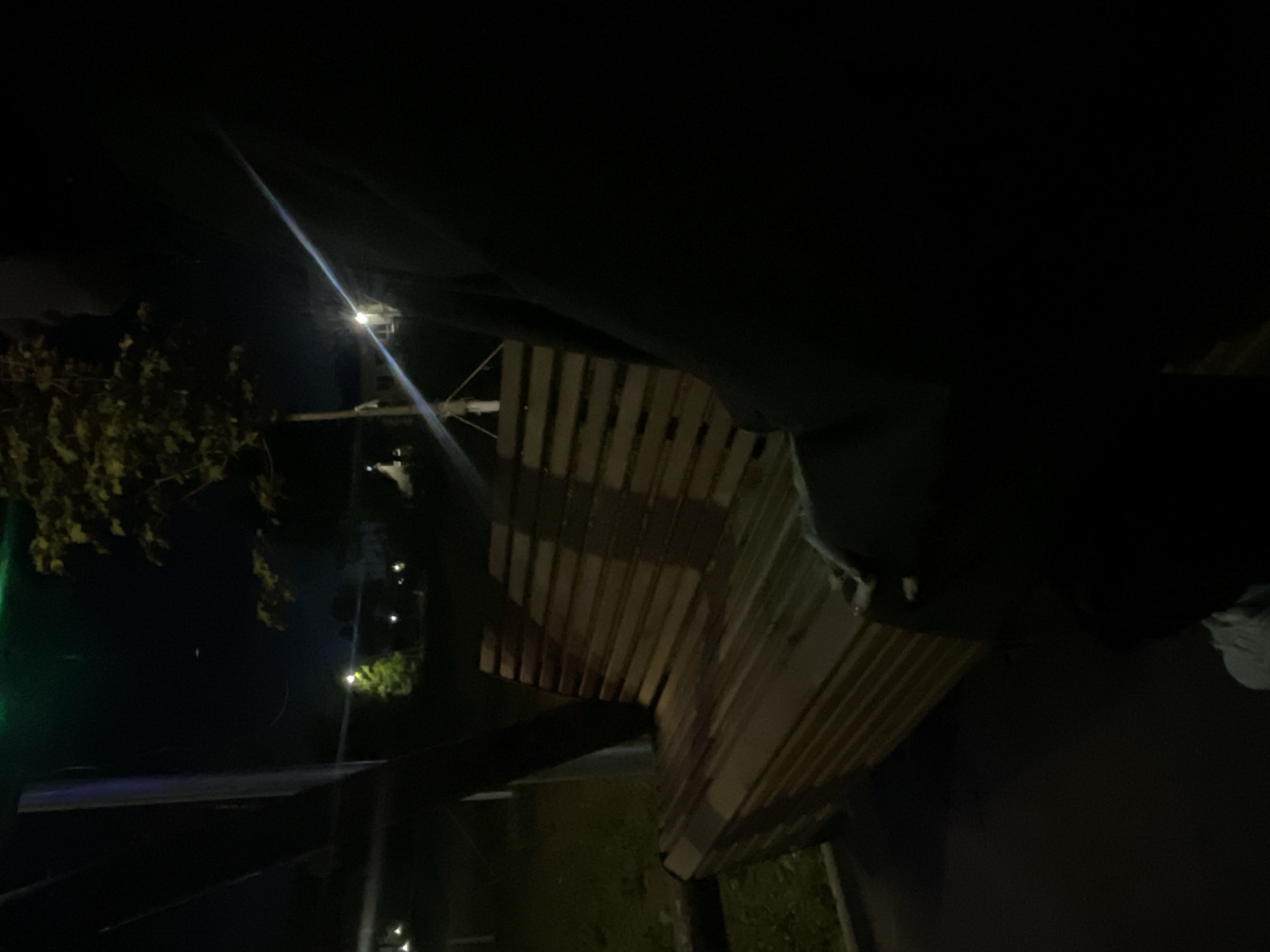 This outdoor photograph, taken at night in Красные Баки, Russia, captures a scene illuminated by artificial lights against a dark background. A prominent wooden structure, likely a pergola, shed, or part of a building's roof, occupies the midground and right side of the frame. It features a distinct slatted or corrugated design, with varying degrees of illumination highlighting its texture and creating shadows.
A bright artificial light source, possibly a streetlamp, is positioned towards the upper-left, emitting a strong lens flare that streaks diagonally across the dark sky. This light illuminates a cluster of green foliage, likely tree leaves or bushes, visible on the left side of the image. Further into the background, other smaller, distant lights can be seen, hinting at a wider illuminated area.
The upper portion of the scene on the right is covered by a large, dark, undefined mass, possibly a tarp, canopy, or an overhang. No individuals or specific activities are visible in the static composition. The overall atmosphere is dark and quiet, with the artificial light creating points of focus and contrasting with the surrounding shadows. There is no visible text.