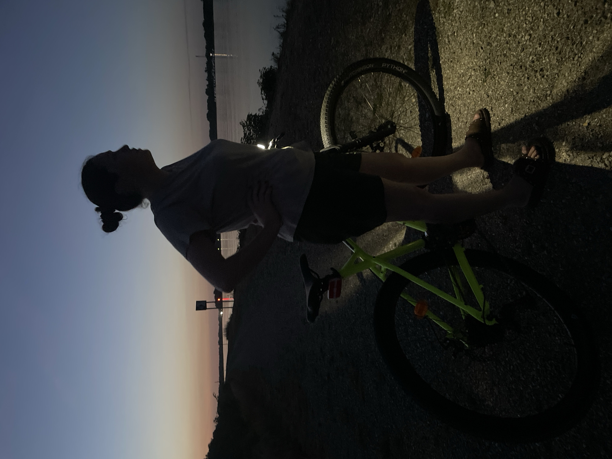 A person stands with a neon green bicycle at dusk on a gravel path next to a body of water. The person is silhouetted against a pale orange and blue sky, with their hair pulled up in a bun. They are wearing a t-shirt and shorts, and sandals. The bicycle has knobby tires, and a red reflector on the back. The water appears to be a river or lake. In the distance, there are lights on the opposite shore, possibly from buildings or boats. A navigational marker is visible in the water. The scene is set at the Rhein River in Ottenheim, Germany.