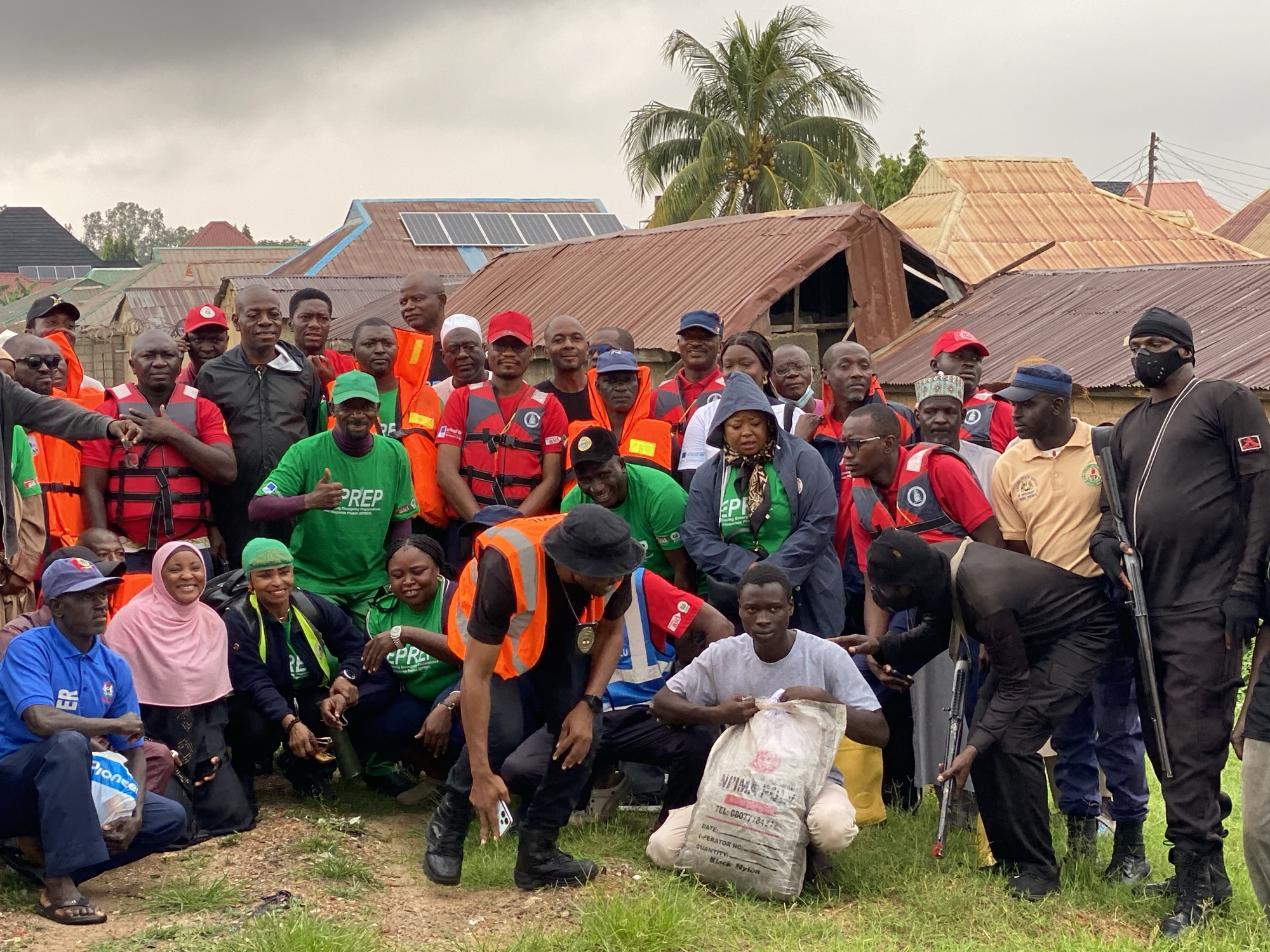 In Kawo, Nigeria, a diverse group of individuals, including men and women, are gathered in a dusty, outdoor setting. The majority are wearing vibrant orange and red life vests over their clothing, suggesting a water-related activity or emergency preparedness. Some are also wearing green shirts with "PREP" printed on them, possibly indicating a preparedness program.
The group consists of people of various ages and ethnicities, with many appearing to be adults. Their expressions range from serious to smiling, and some are interacting with each other. Several individuals are kneeling or crouching, while others are standing. One man in the foreground is holding a large, opaque bag that appears to contain a dry substance. Another person in an orange vest is holding a smartphone.
The background reveals a neighborhood with a cluster of houses, many with corrugated metal roofs. Some rooftops are equipped with solar panels. A tall palm tree is visible in the distance, characteristic of the tropical climate. The overall atmosphere suggests a community event or a response to a local situation.