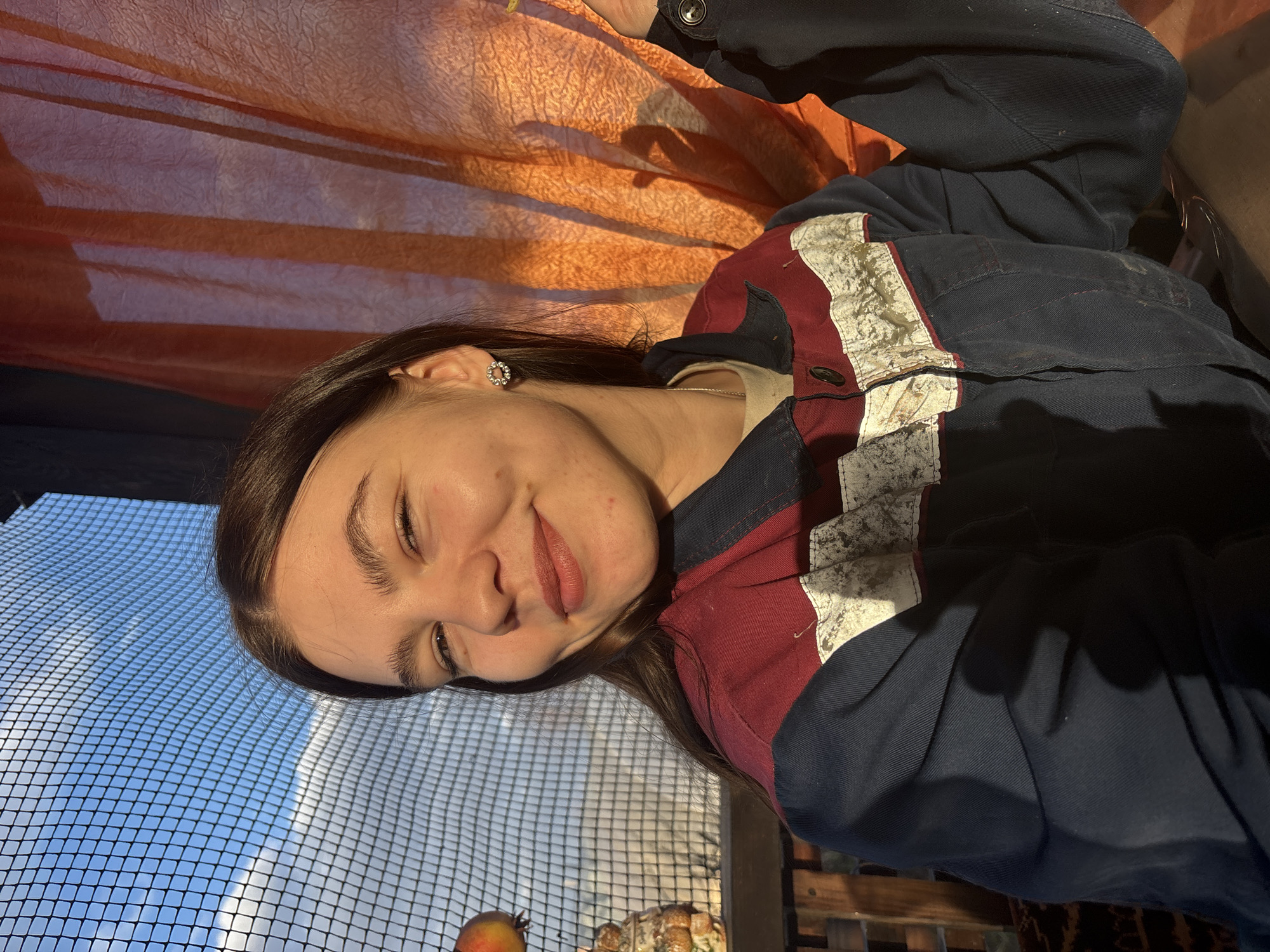 A young woman with long brown hair is indoors, possibly in Chaikovsky, Russia, lying on a wire mesh surface with visible sky and sunlight. She is wearing a navy blue work jacket with red and white striped detail. The background includes an orange curtain. An apple and an indiscernible dish are also visible.