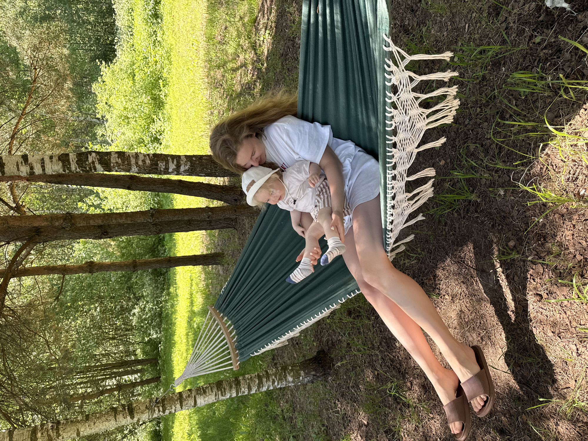An adult female and an infant are situated within a dark green fabric hammock suspended between two trees in Копнино, Russia. The hammock features white macrame-style fringe and white rope suspensions, anchored to a birch tree on the left and a darker-barked tree on the right. The female, with long light brown hair, wears a white short-sleeved t-shirt, white shorts, and brown slide sandals. She is seated, embracing the infant with her left arm, her right hand holding the infant's leg. Her head is tilted towards the infant. The infant wears a light-colored t-shirt, light-striped shorts, a white bucket hat with "BROWN" text, and blue and white striped socks. The infant is oriented towards the female. The ground beneath the hammock is covered with dry leaves, pine needles, and sparse green grass, with brighter green grass visible in the sunlit mid-ground. A dense forest with varied deciduous and coniferous trees forms the background under bright daylight, casting distinct shadows.