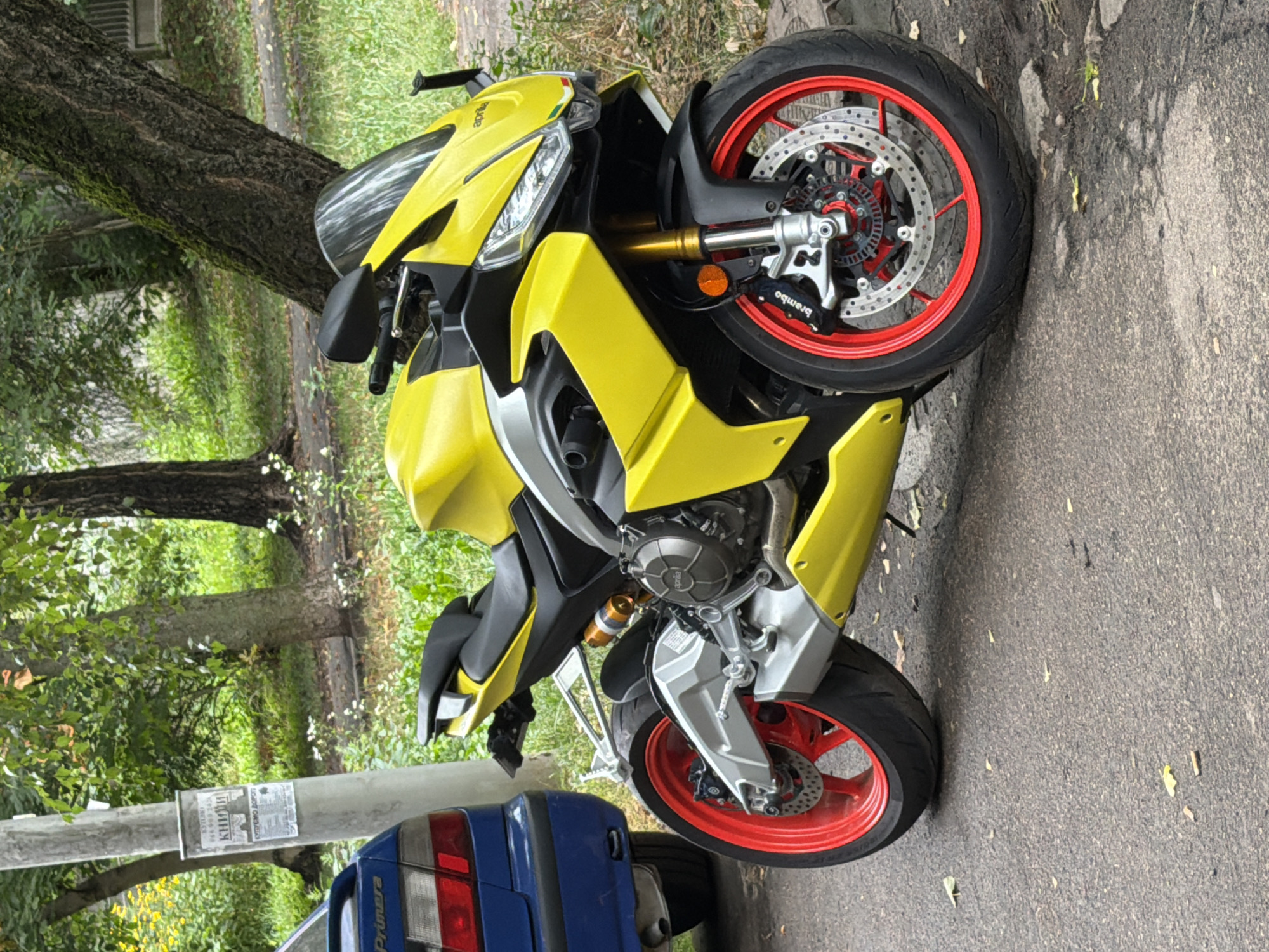A bright yellow sport motorcycle is parked on a paved street next to a blue sedan. The motorcycle has red rims and silver accents. A large tree with mossy bark is behind the motorcycle, and lush green foliage is visible in the background. A utility pole with a poster is to the left of the motorcycle. The setting appears to be a residential area in Kyiv, Ukraine, based on the Cyrillic text on the poster. The scene is static, with no people or ongoing actions depicted.