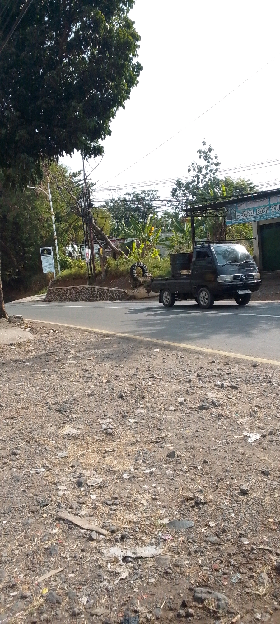 A black utility vehicle is driving down a paved road. The vehicle is traveling from left to right, and it is moving away from the camera. The road is bordered by a dry, rocky embankment on the left and a grassy shoulder on the right. In the background, there are trees, utility poles, and buildings. One of the buildings has a sign that reads "JUAL BAN VULC," which translates to "FOR SALE VULCANIZED TIRES." This indicates the image was likely taken near a tire shop in Cilaku, Indonesia.