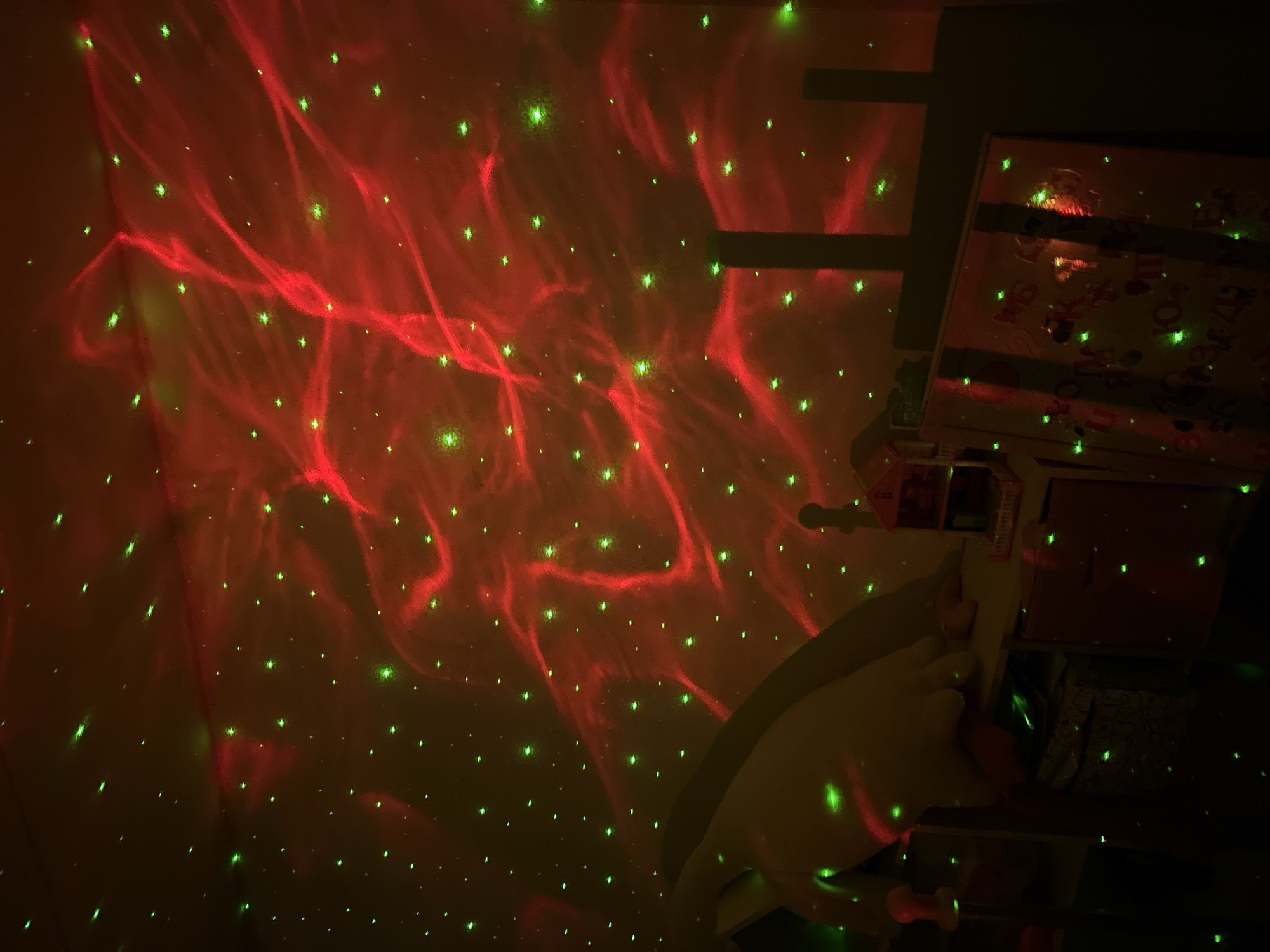 The image appears to be taken indoors, likely in a bedroom or playroom. The wall is illuminated by a light projector displaying a pattern of red nebulas and green stars. In the right portion of the image is a structure that seems to be a toy dollhouse with multiple levels, drawers, and items within it, also illuminated by the light projector. A white object, possibly a pillow, is visible near the base of the dollhouse. The scene has a dark and moody atmosphere due to the lighting conditions.
There is no evidence to suggest this is in the city of Chaikovsky, Russia.