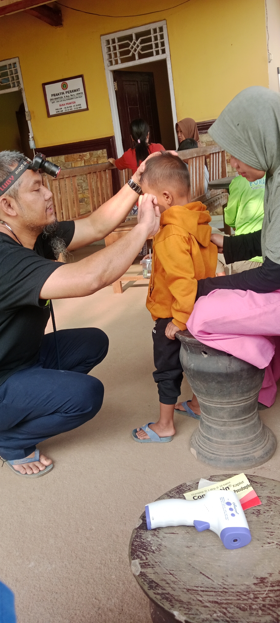In the city of Sidodadi, Indonesia, a medical professional wearing a headlamp is examining a young child's eye. The child is wearing an orange hooded sweatshirt and dark pants, and is being held steady by another individual, possibly a parent. A woman in a grey hijab is seated nearby. The examination is taking place outdoors, in front of a building with yellow walls and a wooden railing. A sign on the wall reads "PRAKTIK PERAWAT" and provides opening hours. A digital thermometer is visible on a wooden table in the foreground. The scene suggests a community health outreach or a clinic operating in a residential area.