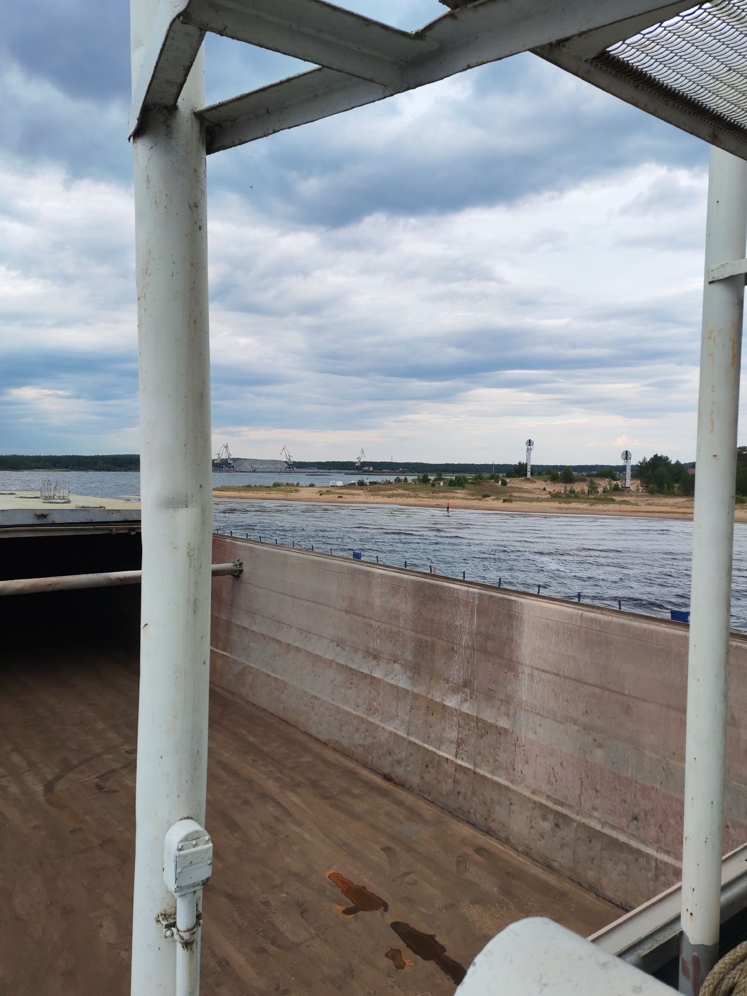 The image depicts a view from a barge on the Shalsky River in Shalsky, Russia. The barge's deck and cargo hold are visible in the foreground, with a large white pipe prominent on the left. The river stretches out towards a sandy bank dotted with trees and two navigation markers. In the distance, industrial cranes and a port facility are visible on the far bank under a cloudy sky. A lone figure is standing on the sandy bank. The overall scene suggests a working waterway and industrial activity in a rural setting.