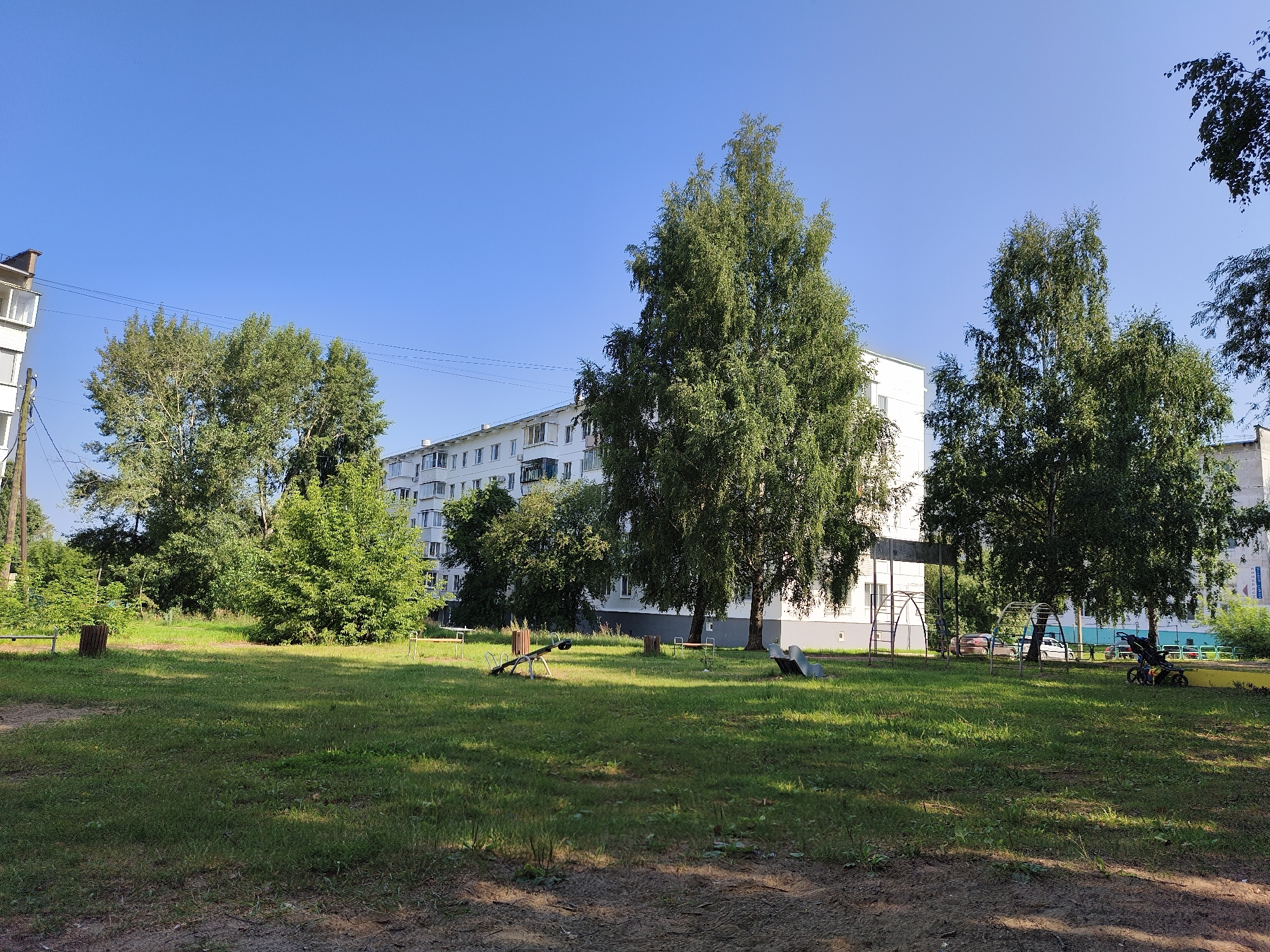 The image depicts a residential courtyard in Chaikovsky, Russia, under a clear blue sky. In the background, a multi-story apartment building with white and grey facades dominates the scene. Several mature trees with dense green foliage are scattered throughout the courtyard, casting shadows on the grassy ground. In the foreground, a patch of dirt and dry leaves is visible. Scattered within the courtyard are elements of a children's playground, including a seesaw, a slide, and a climbing structure. Two benches, one wooden and one metallic, are also present. A trash bin made of wood is situated near one of the benches. In the middle ground, a double stroller is parked. The overall impression is of a quiet, everyday urban setting during daylight hours.