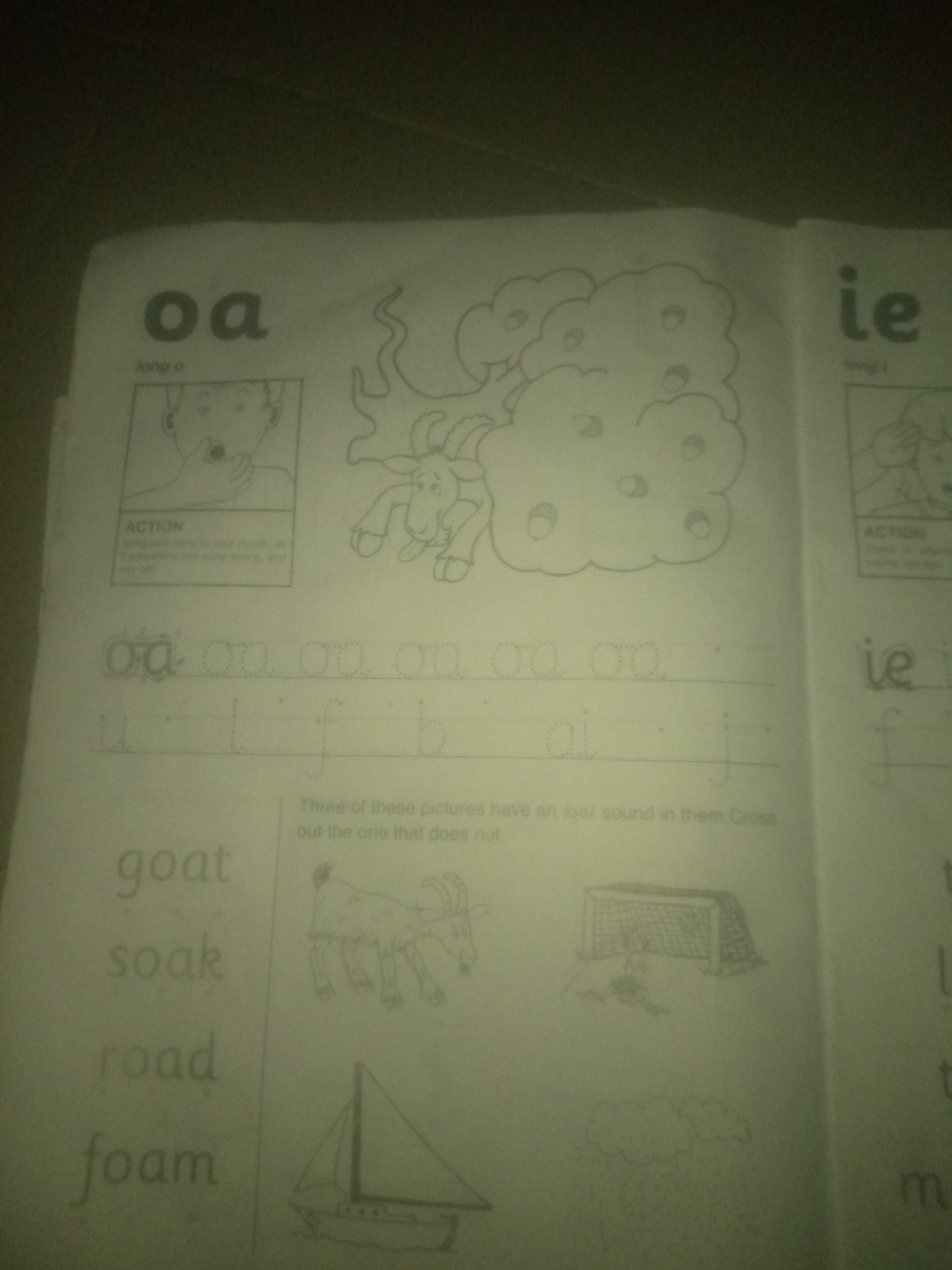 The image shows a page from a children's educational workbook focusing on phonics. On the left side, the digraph "oa" is displayed prominently, followed by the description "long o" and an illustration of a mouth making the "oa" sound. Below this, there are practice lines for tracing the digraphs "oa" and "ai". Further down, words like "goat", "soak", "road", and "foam" are presented in large print.
To the right of the "oa" section, there's a drawing of a goat in front of a cloud, likely illustrating the "oa" sound in "goat".
On the right side of the page, the digraph "ie" is shown, with a similar "ACTION" box and practice lines. Below this, there are some unclear words or letters and an illustration of a soccer goal with a player.
The bottom part of the page contains drawings of a sailboat and clouds, also likely related to the phonics exercises.
The image is a close-up, angled shot of the workbook. The lighting is uneven, casting shadows and making some parts of the page appear lighter or darker. The background is dark and out of focus. There is no indication of the city of Jauro Yinu, Nigeria in the image.