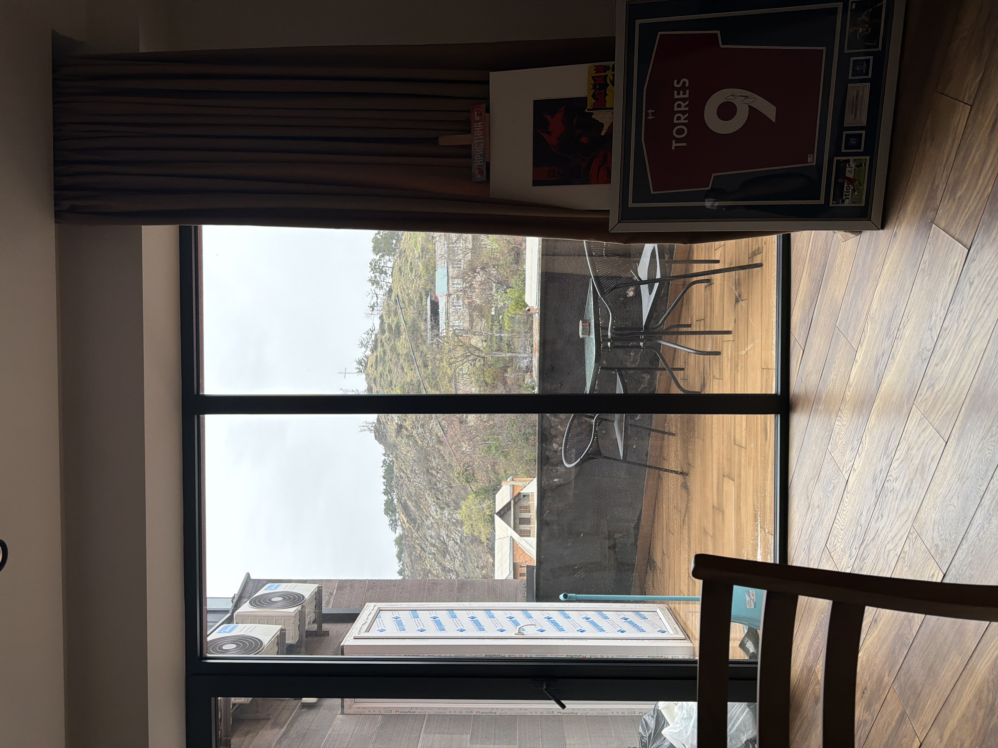 The image displays an interior view looking outwards through large, multi-pane glass doors onto an outdoor balcony and a distant hillside under an overcast sky. The location is Tbilisi, Georgia, specifically indicated by the prominent cross structure visible atop the distant hill, consistent with Mtatsminda.
In the foreground inside, a wooden plank floor occupies the lower portion of the frame, with the partial back of a wooden chair visible on the left. To the right, a framed red football jersey, displaying "TORRES" and the number "9," leans against an unseen surface on the floor. Adjacent to it, an easel holds a white canvas featuring a dark, stylized painting of Batman with red accents. Above this artwork, a smaller item with multiple Batman logos is present, and on top of the easel, a small sign reading "КРИСТИНА" (Kristina) in Cyrillic script is placed. A brown, pleated curtain is fully drawn on the right side of the glass doors.
The outdoor balcony features wooden decking, which appears damp in various spots. A small, round, dark-colored outdoor table is set with two matching chairs. A low, dark-colored concrete or stone wall forms the balcony's railing. To the left, a partially open white door, covered with protective blue and white plastic film (indicating renovation or new installation), provides access to an adjacent area. Two external air conditioning units are mounted on the wall to the left of this door, and several white plastic bags are visible on the ground. A blue vertical pipe is also present near the left edge of the balcony.
Beyond the balcony, a steep, rocky hillside covered with sparse vegetation, including deciduous trees and bushes, rises under a uniformly grey, overcast sky. Scattered residential buildings with pitched roofs are visible on the hillside. The distinct cross structure is clearly discernible at the summit of the hill. No people are visible in the scene.