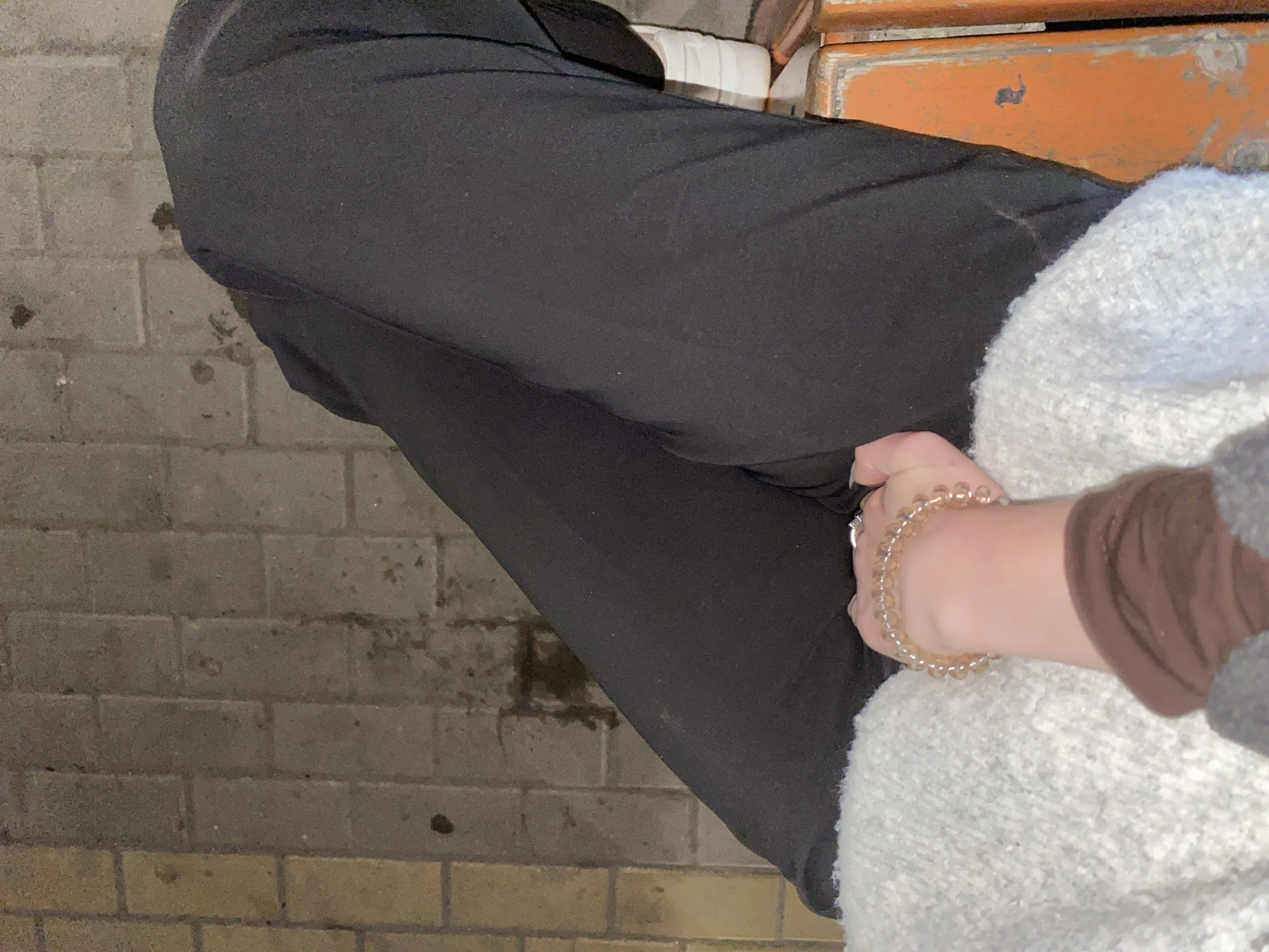 The image captures a close-up, low-angle view of a person seated or resting, likely in an outdoor or semi-outdoor setting. The subject's torso is partially obscured, but their legs are prominent, clad in black trousers. A hand, adorned with a light-colored, translucent beaded bracelet, rests on their lap, which is covered by a light-colored, possibly white or light grey, fluffy or knitted garment. A brown long-sleeved top is visible on the arm.
The ground beneath the person is paved with rectangular grey bricks or pavers, showing some dark stains or wet patches. In the upper right corner, an orange-brown structure, possibly a weathered wooden or metal bench, is visible, partially covered by the person's upper body. The lighting is dim, suggesting either an indoor setting with low illumination or an outdoor scene during dusk or nighttime. No specific activity beyond sitting or resting is discernible. No text is visible in the image.
