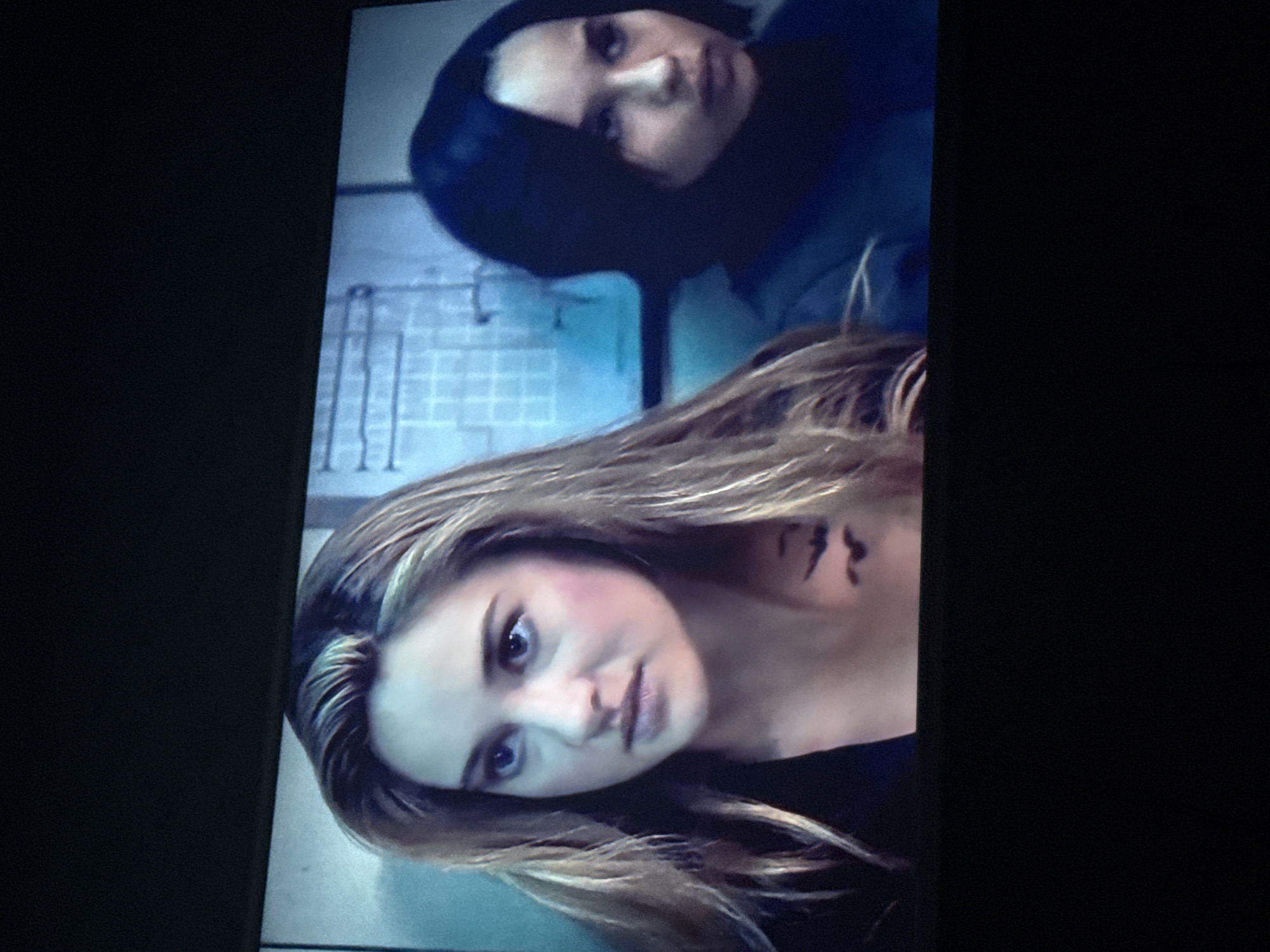 The image shows two women. One woman has long blond hair and a tattoo on her neck. The other woman has short black hair and is wearing a dark hooded jacket. The background is a light blue wall with a pattern of squares. There is no information to indicate the location in Mytishchi, Russia.