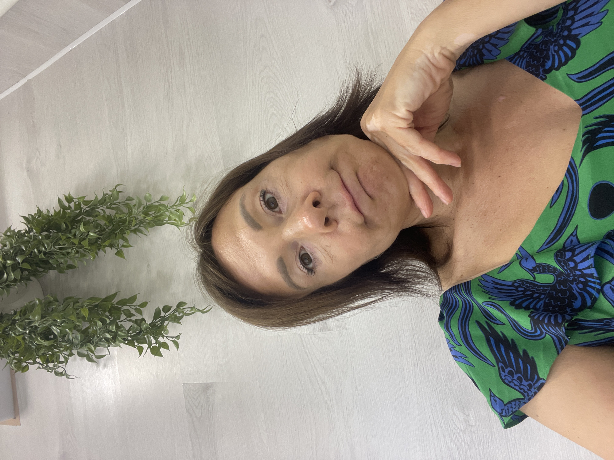 The image shows a woman posing for a selfie. She is wearing a green dress with a blue bird pattern. She has her right hand on her chin and is looking at the camera with a playful expression. The background is a light-colored wooden floor and some green plants. The image appears to be taken indoors during the day. The woman has fair skin and brown hair. There are no visible text. The location context is Sochi, Russia.