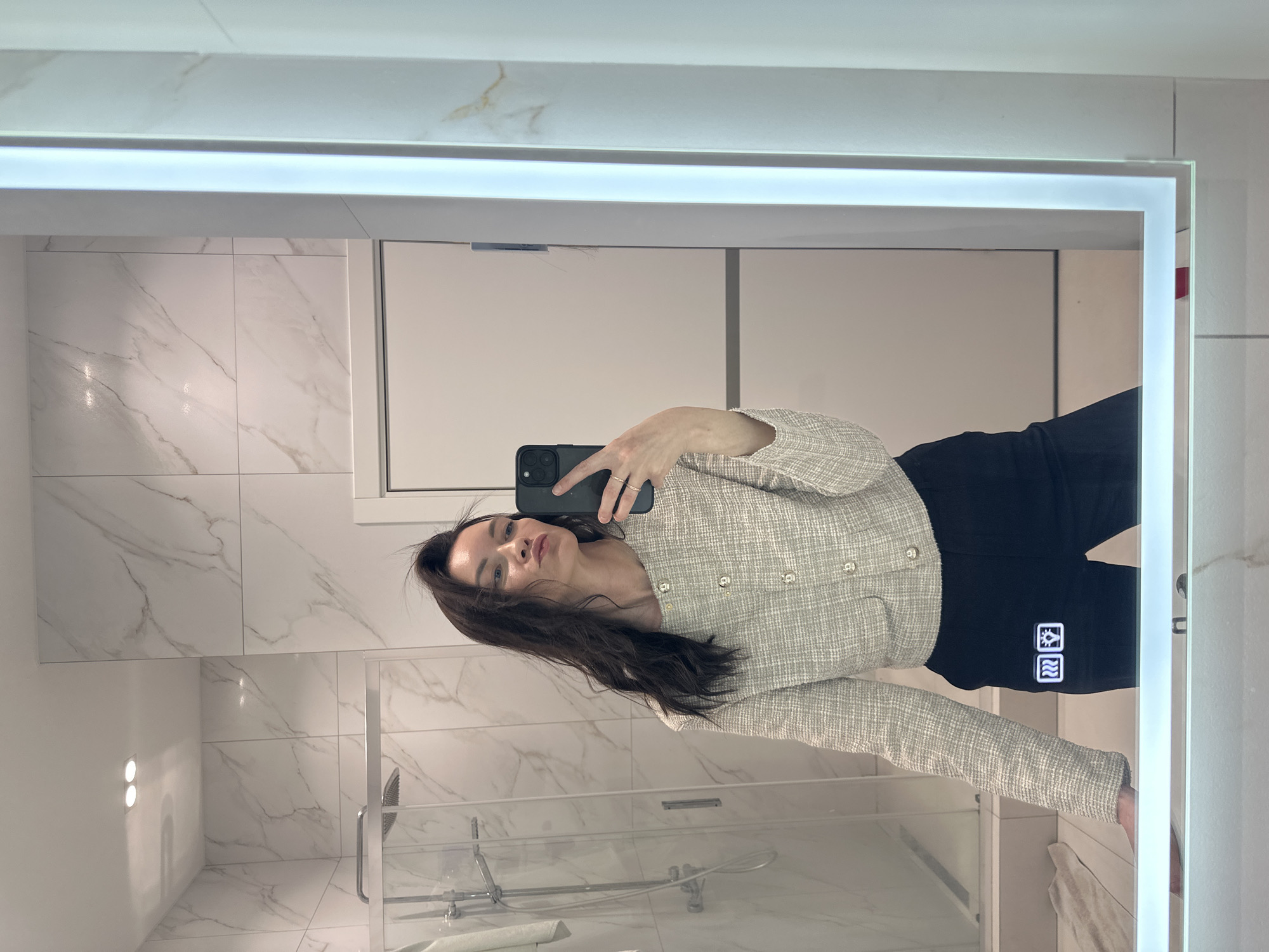 A woman is taking a selfie in a bathroom mirror. The bathroom has white marble-patterned tiles on the walls and a glass shower door is visible. The woman is wearing a light-colored textured jacket and black pants. The location is unknown.